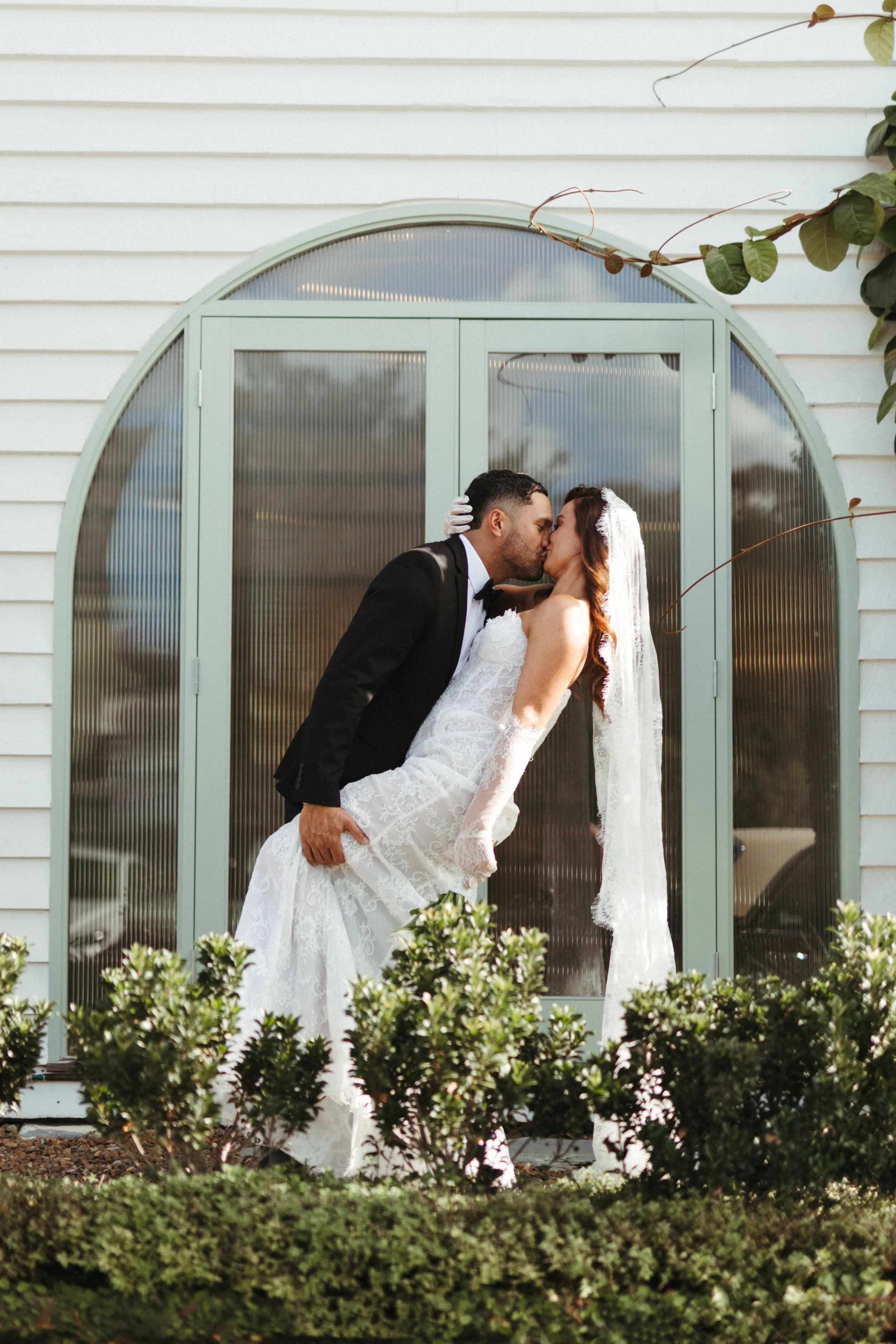 A couple in wedding attire sharing a kiss in front of a pale green arched window on a white house, with greenery in the foreground.