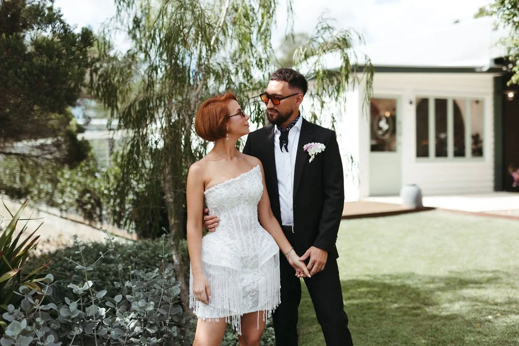A woman in a white lace dress and a man in a black suit with a boutonniere, standing outdoors, holding hands, under a tree near a house.
