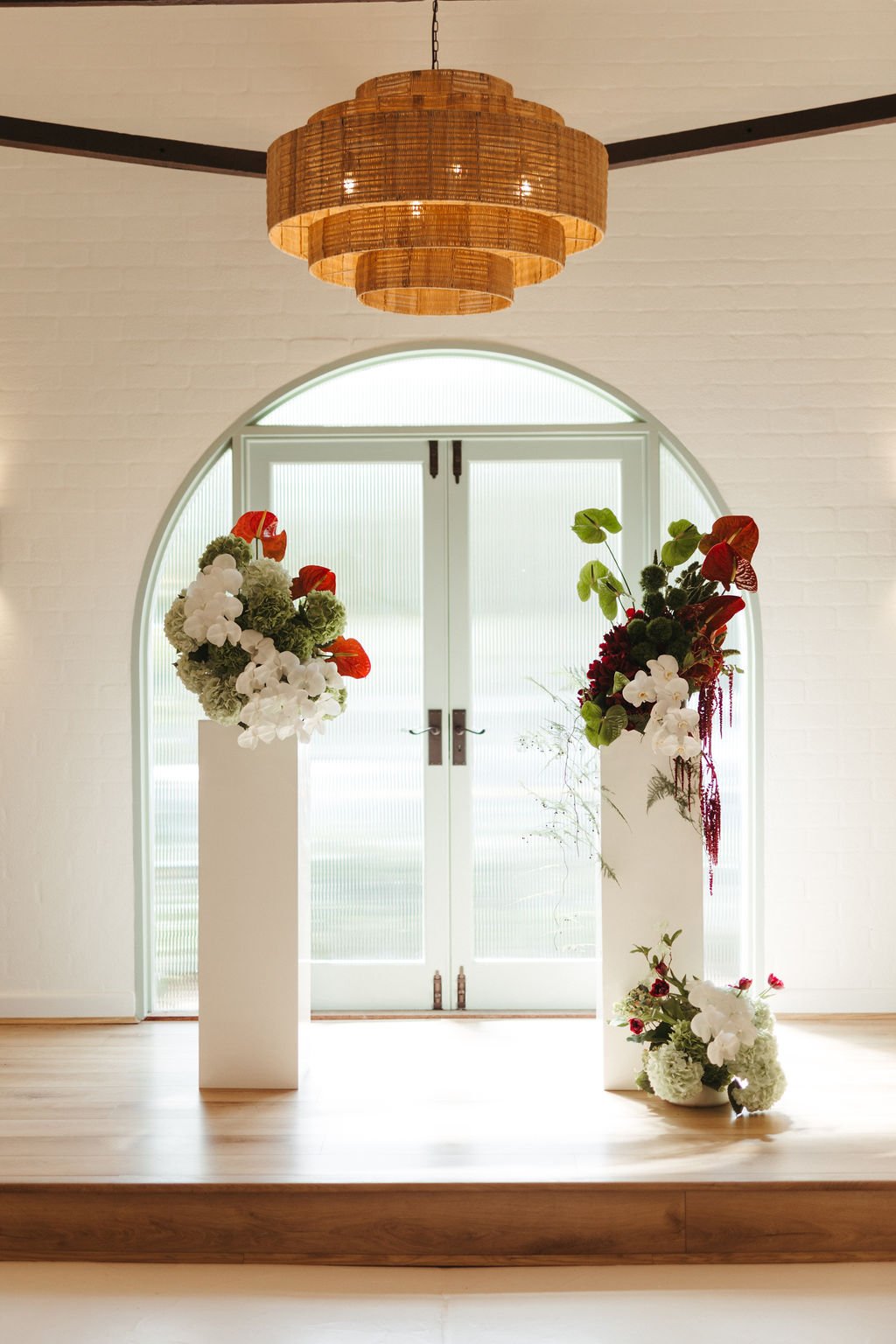 Two tall white vases filled with floral arrangements, placed on a wooden platform in front of glass double doors with frosted glass, and a woven wooden chandelier hanging from the ceiling.