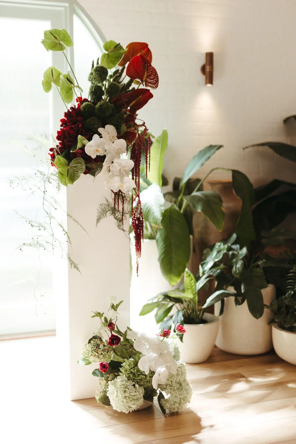 Indoor floral arrangement with orchids, roses, and greenery near a window, with numerous potted houseplants on a wooden floor.