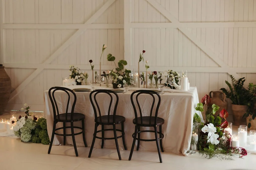 A rectangular dining table with a cream-colored tablecloth, decorated with candles and floral arrangements, situated in a white-walled barn interior, surrounded by black chairs and potted plants.