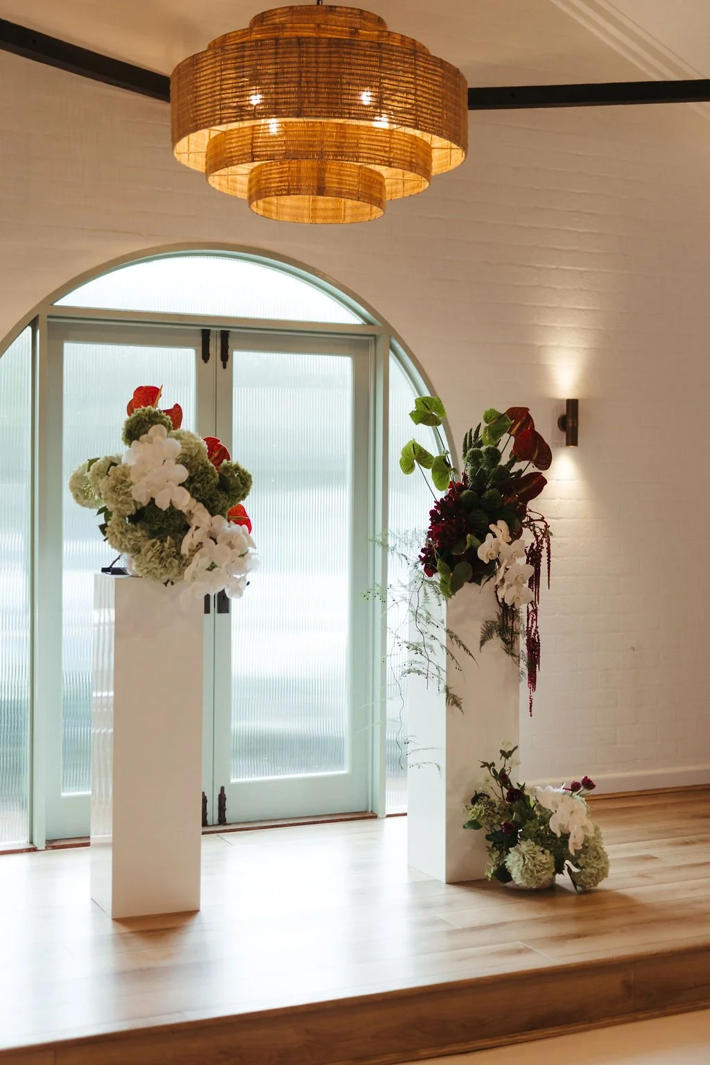 Elegant interior with white walls, arched frosted glass double doors, hanging wicker light fixture, and floral arrangements on white pedestals in a well-lit room.