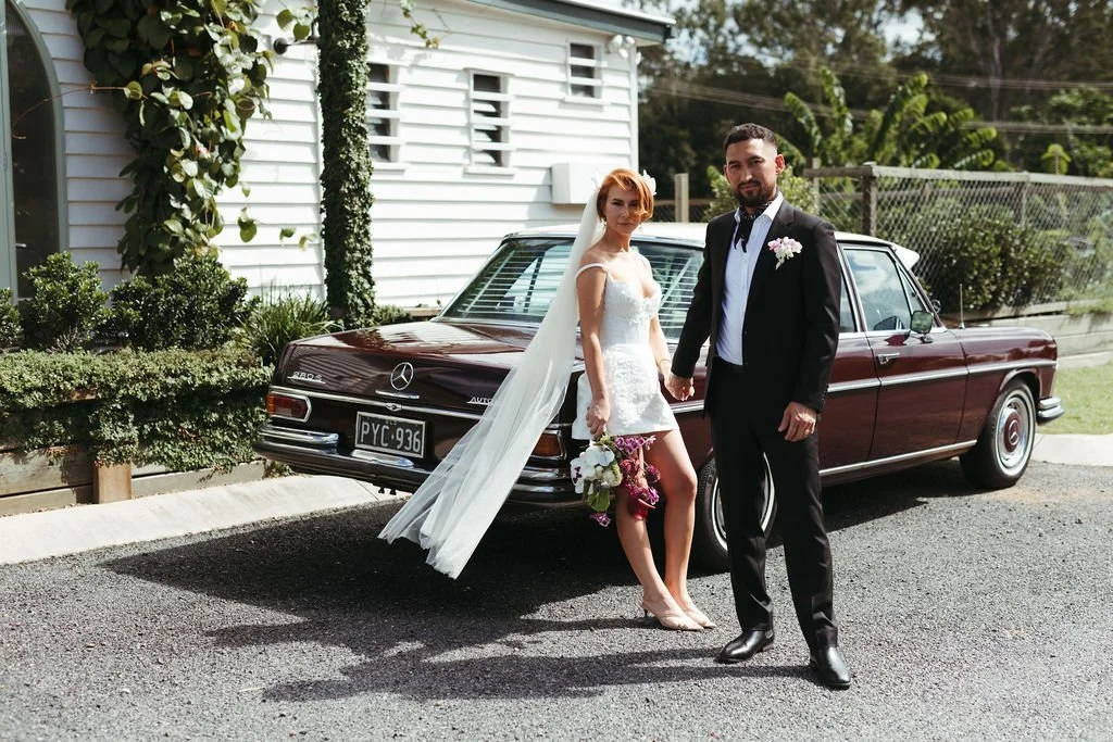 A bride and groom stand holding hands in front of a vintage Mercedes-Benz car outdoors on a driveway. The bride wears a white wedding dress with a veil and holding a bouquet. The groom wears a black tuxedo with a boutonniere. A white house with plant