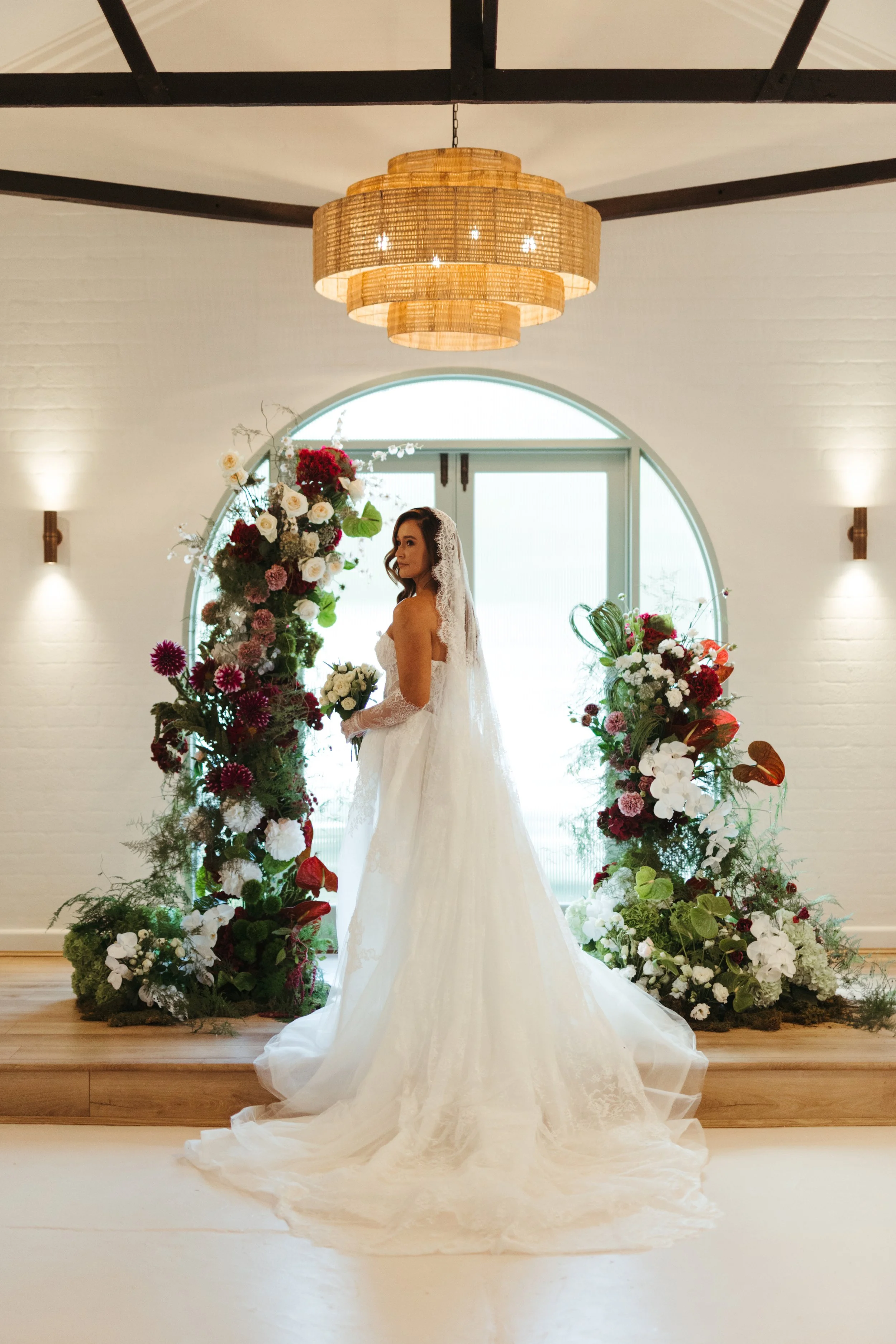 A bride in a white wedding gown with a long train and veil standing in front of a large window. She is surrounded by tall floral arrangements with various flowers including roses and orchids. The setting appears to be indoors with a wooden floor, whi
