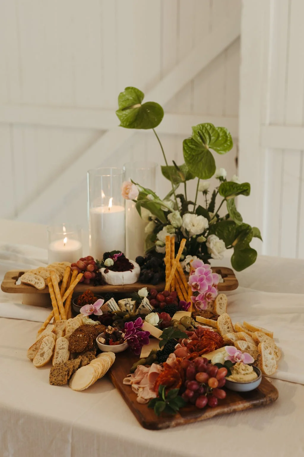 A charcuterie board with cheese, grapes, crackers, and meats, decorated with pink flowers, on a table with white candles and a floral centerpiece against a white paneled wall.