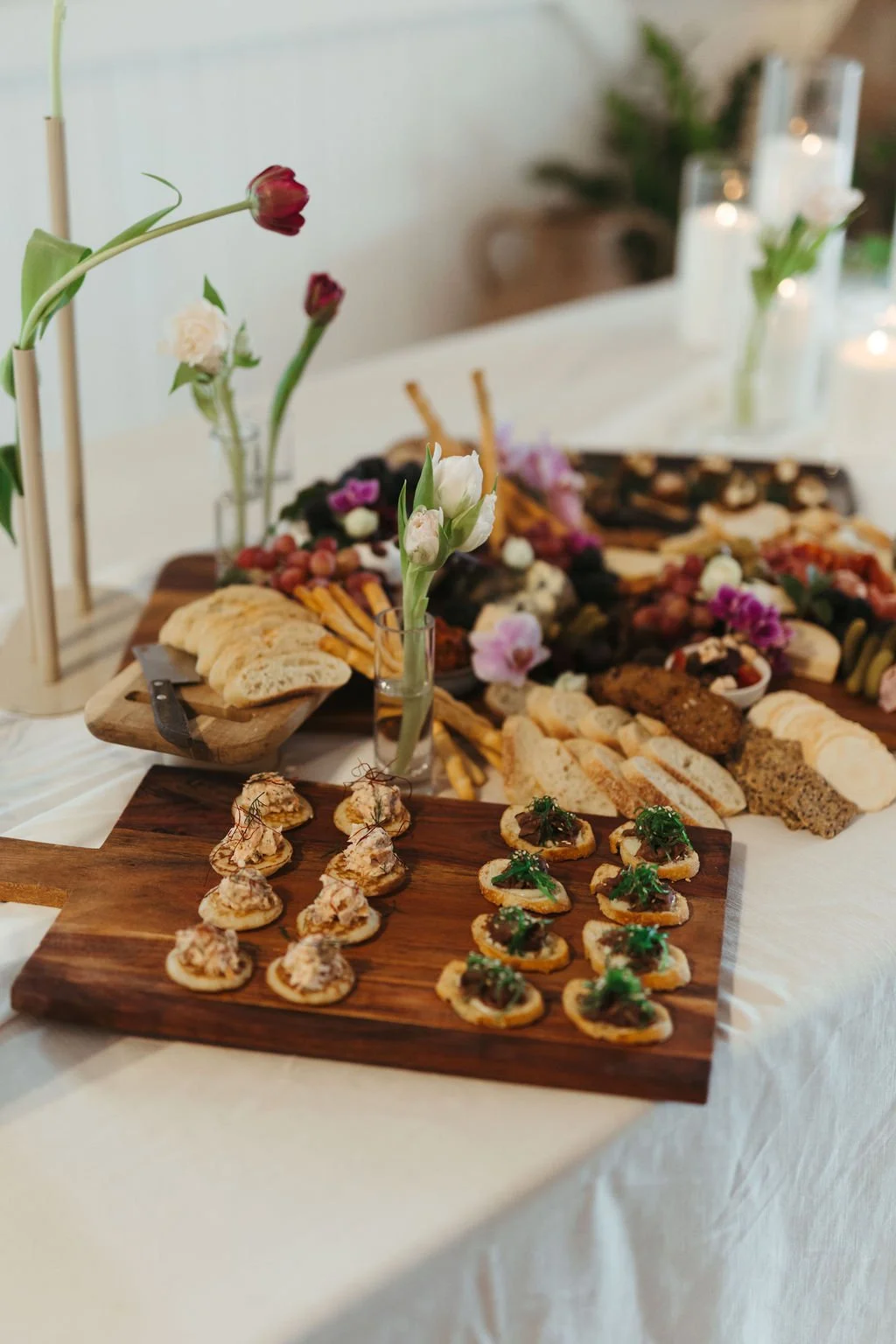Elegant spread of appetizers and finger foods on wooden boards and trays, decorated with vases of flowers, set on a white tablecloth.