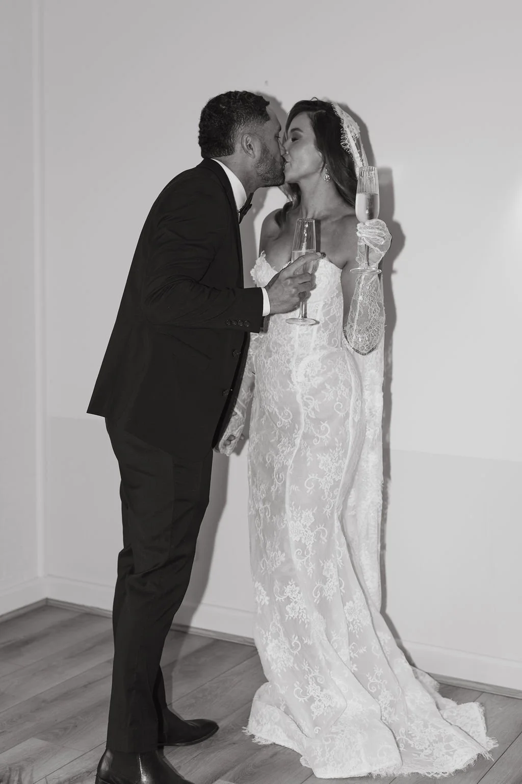 A black and white photo of a bride and groom kissing while holding champagne glasses; the bride is wearing a lace wedding dress and veil, and the groom is in a tuxedo.