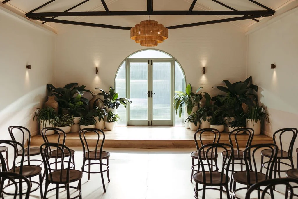 Indoor space with a small stage decorated with large potted plants, a glass door with an arched window, and chairs arranged in front, decorated with small wall sconces and a woven ceiling light.