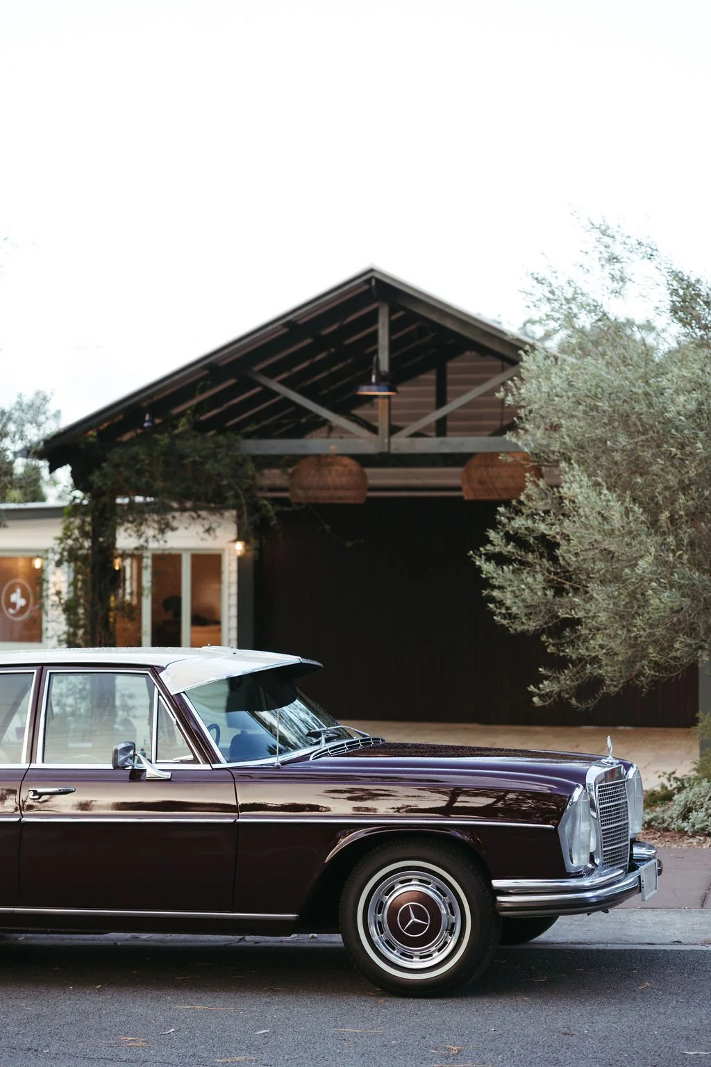 A vintage black Mercedes-Benz parked in front of a modern building with a black and wood exterior, surrounded by trees.