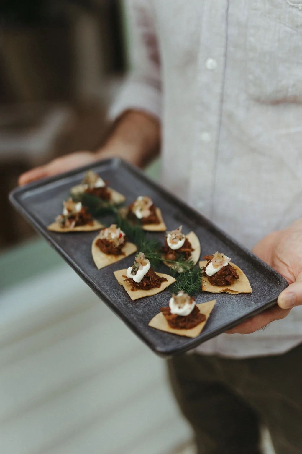 Person holding a black rectangular serving tray with small appetizers on it, each featuring a triangle-shaped chip topped with meat and a dollop of sauce or cream, with a green garnish in the middle.