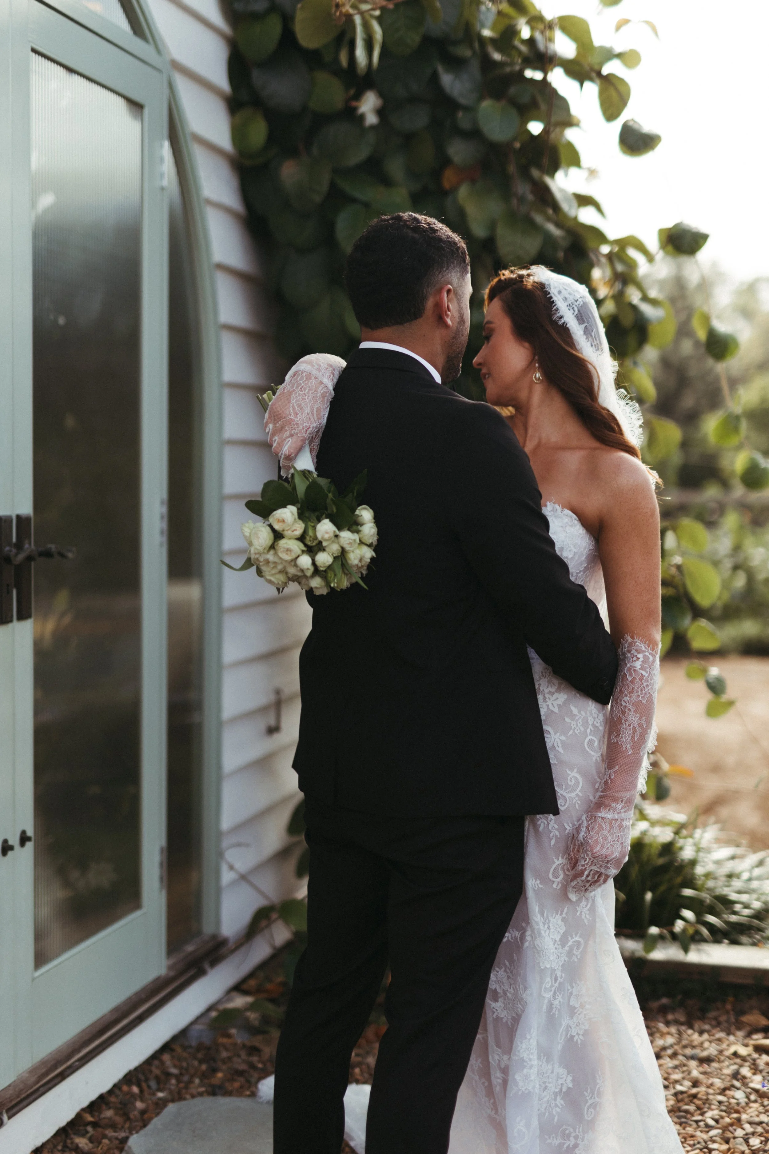 A bride and groom are embracing each other outdoors during sunset, with the bride wearing a lace wedding gown and veil, and holding a bouquet of white roses, standing next to a house with white siding and a large green-leafed vine.