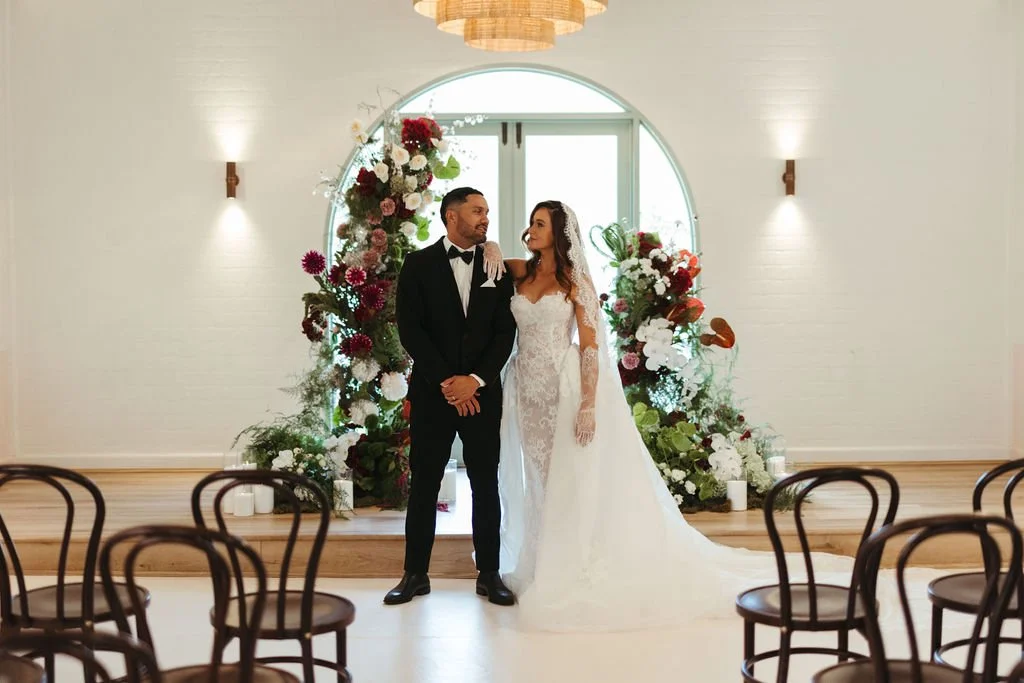 A bride and groom stand together in front of a floral backdrop during their wedding ceremony, with empty chairs in the foreground and a bright window behind them.