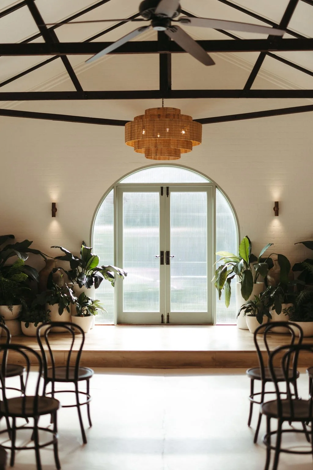 Interior view of a room with a large arched window, potted plants, wooden chairs, and a ceiling fan with a woven light fixture.