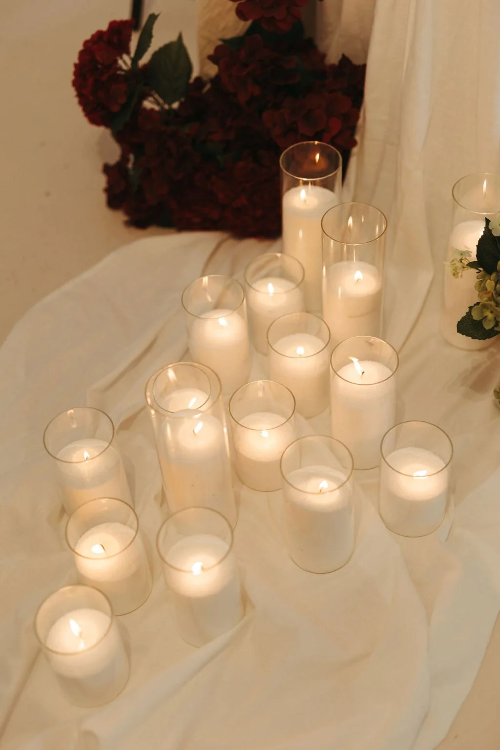 A collection of lit white candles in glass holders arranged on a white cloth with red flowers and greenery in the background.