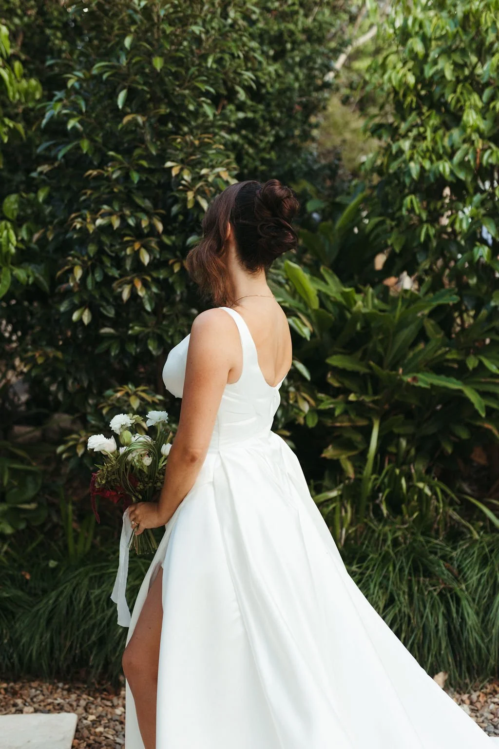 A woman in a white wedding dress holding a bouquet stands outdoors among green foliage, with her back to the camera.