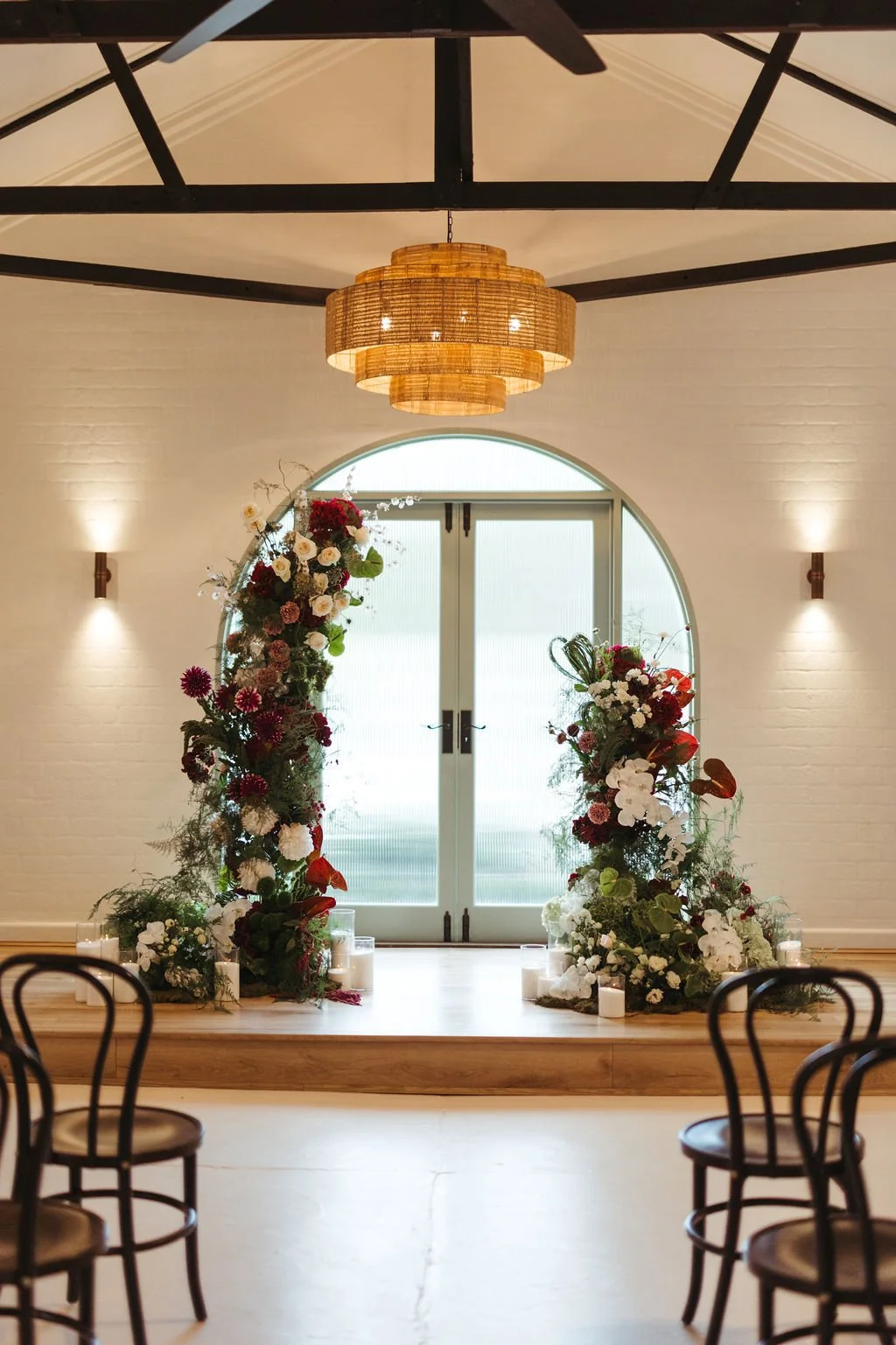 Indoor wedding or event setup with a flower arch, candles, a large window, and a wooden ceiling with hanging light fixture.