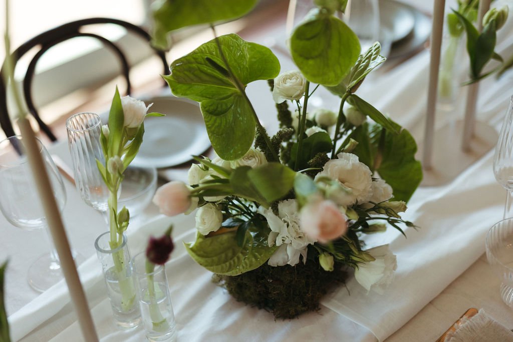 A floral centerpiece with white flowers and green leaves on a table with glasses and a white tablecloth, in a well-lit room.