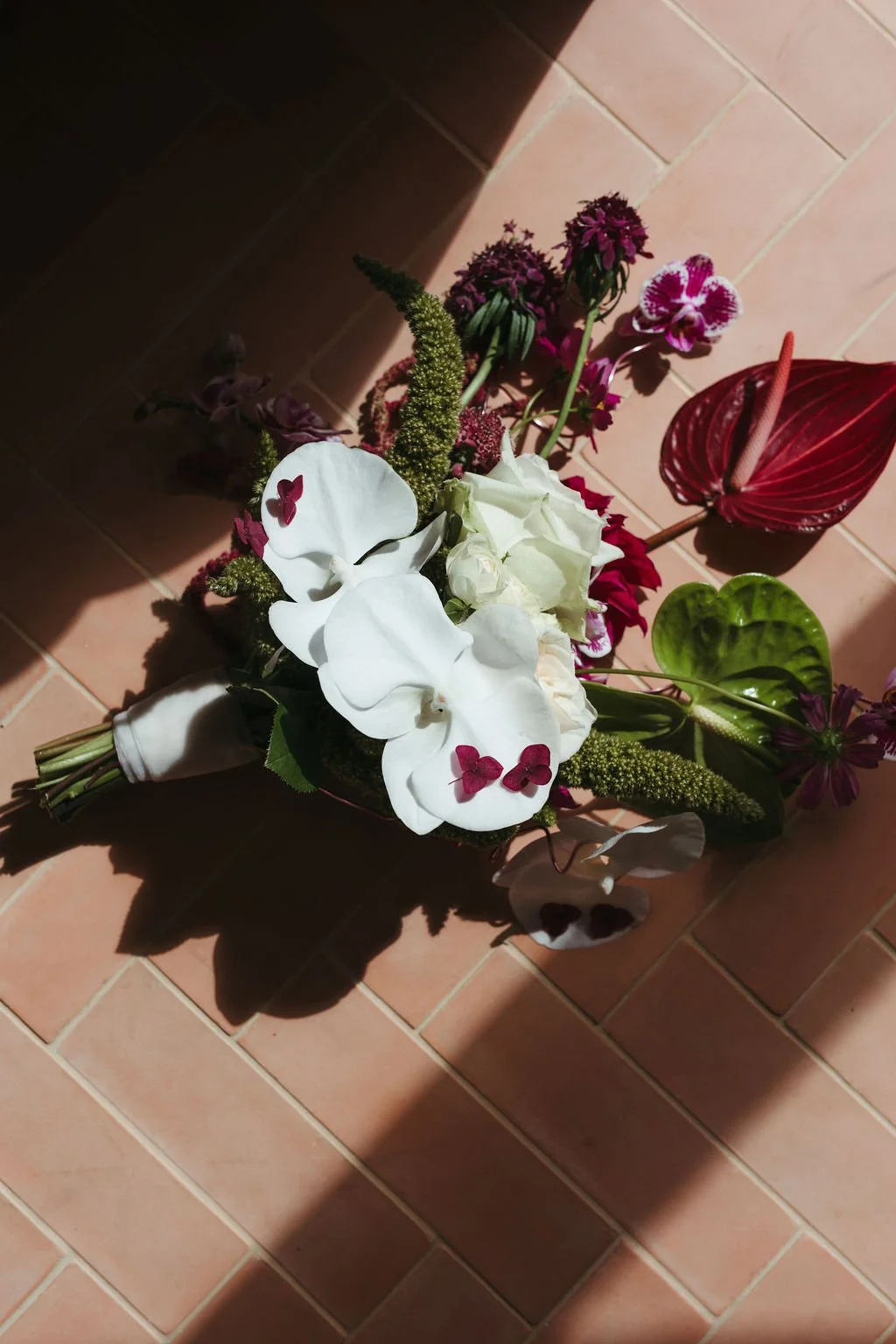 A bouquet of various flowers including white orchids, white roses, and pink and purple blooms, placed on a tiled floor with a shadow cast over part of the arrangement.