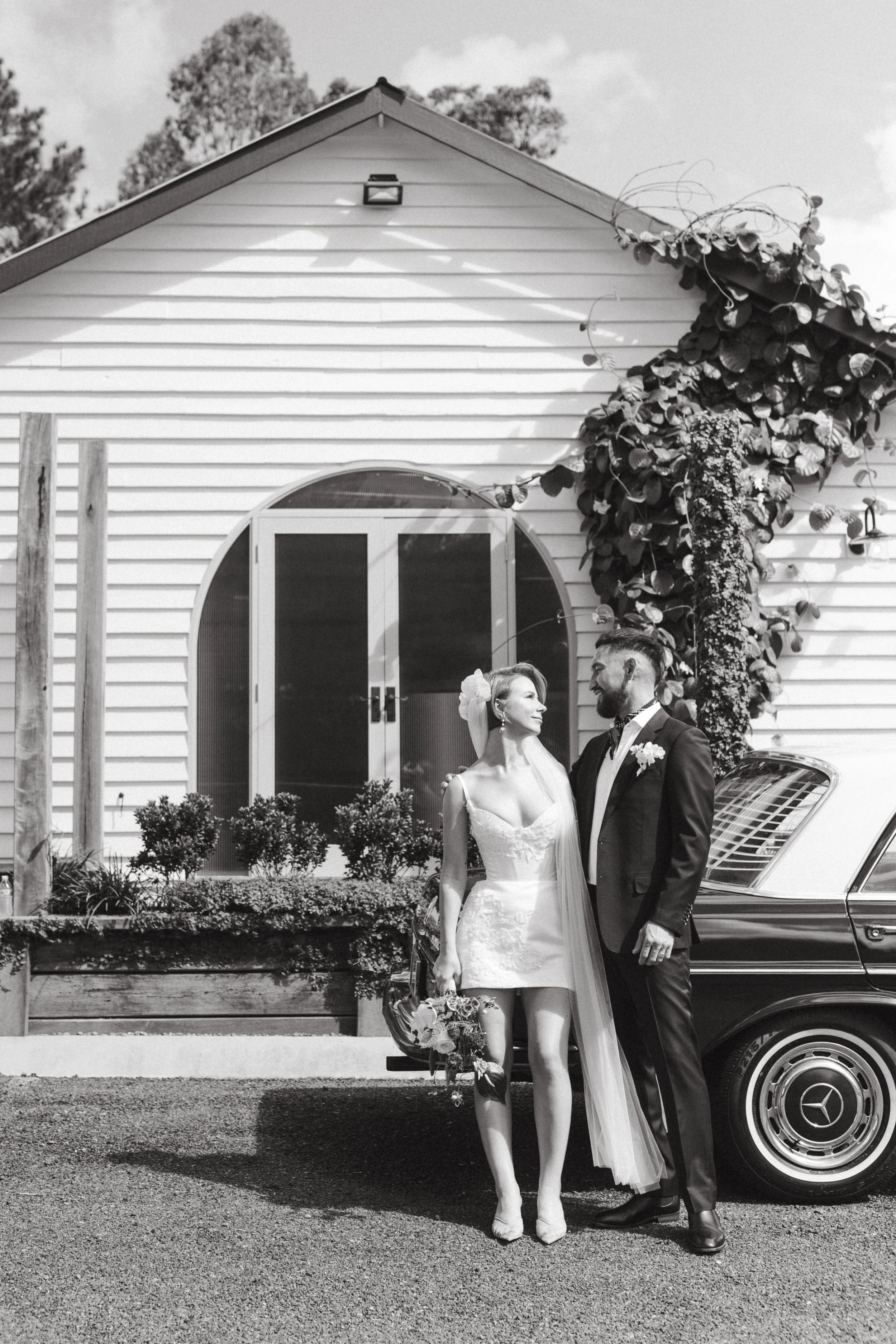 A bride and groom are standing together outdoors in front of a building with a vine-covered wall. The bride is in a white wedding dress wearing heels and holding a bouquet, while the groom is in a tuxedo. They are smiling at each other.