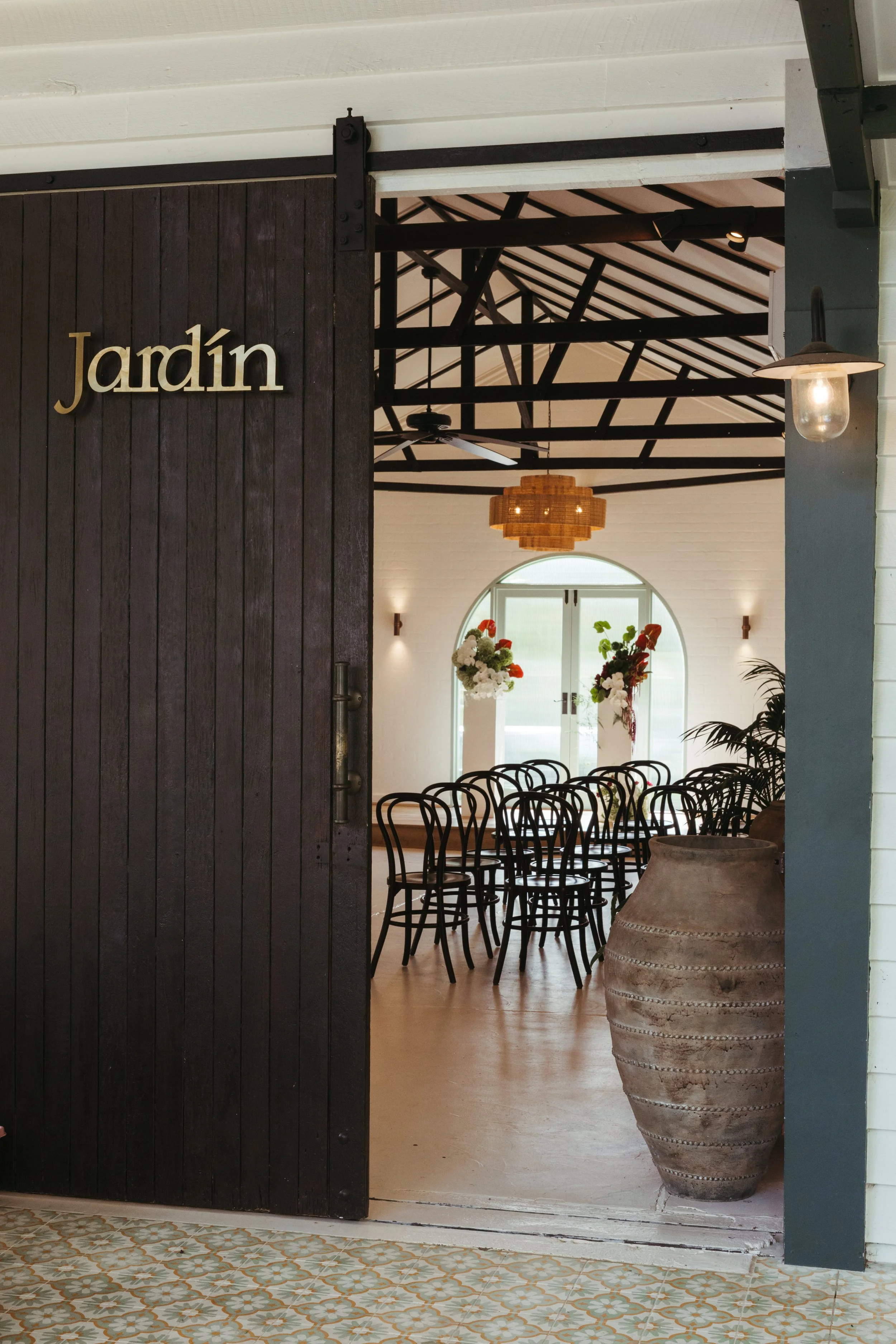 Interior of a dining space with black chairs, floral arrangements, large clay vases, and wooden pendant lighting, viewed through an arched doorway.