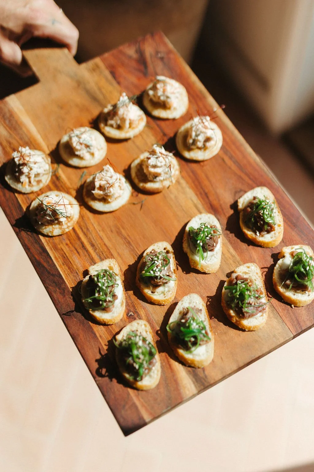 A wooden serving board with two types of small, round appetizers: one topped with smoked fish and microgreens, the other with cooked meat, herbs, and sesame seeds.