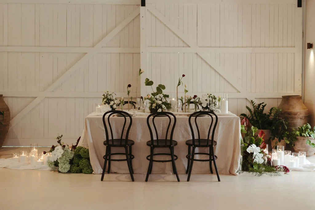 A decorated dining table with a beige tablecloth, black chairs, floral centerpieces, candles, and greenery, set against a white wooden wall with candles and plants on the floor.