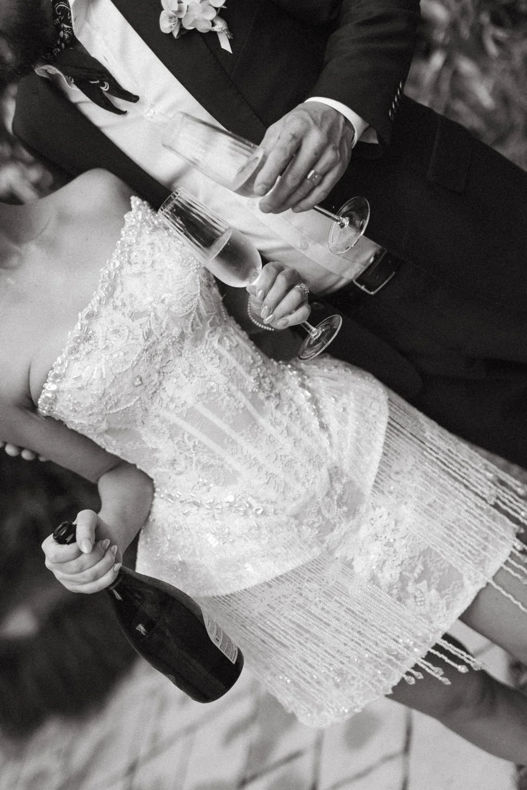 A bride and groom celebrate their wedding with drinks; the bride holds a champagne glass and a bottle, and the groom holds a champagne glass and a bottle.