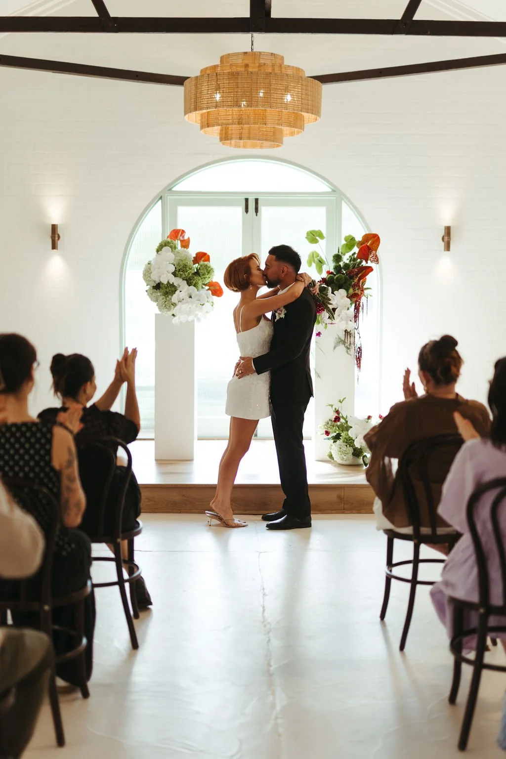 A couple is kissing during a wedding ceremony in front of a window with floral decorations, surrounded by seated guests.