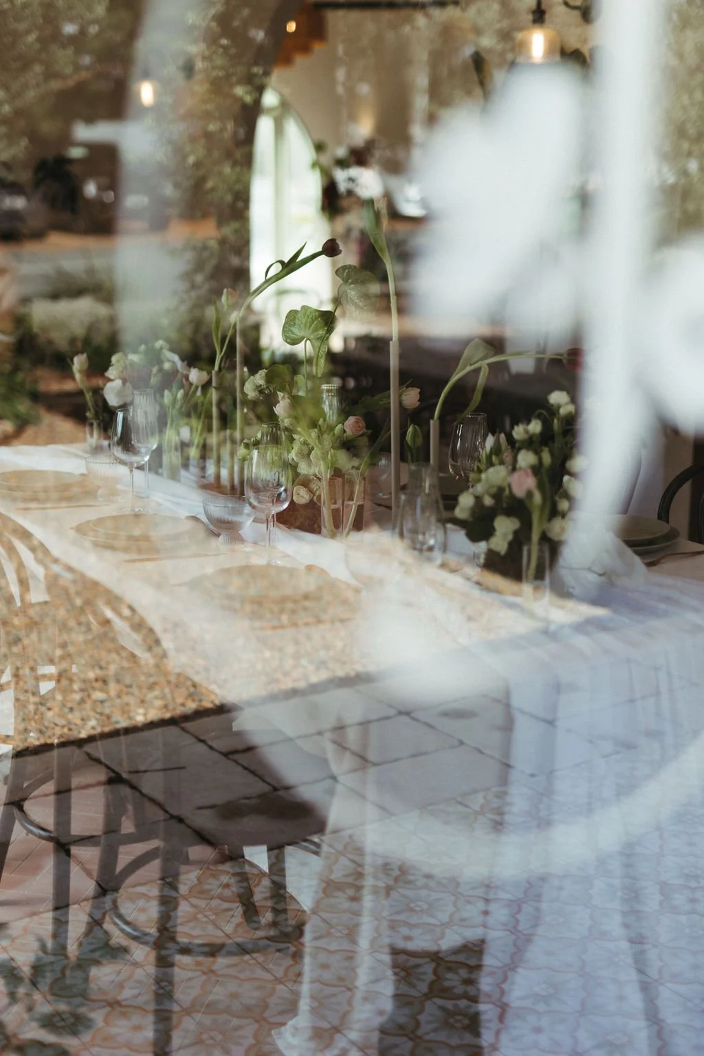 A table set with floral arrangements and glassware, viewed through a window with reflections of the street outside.