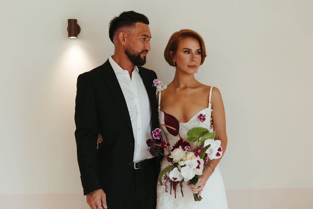 A bride and groom standing side by side indoors. The bride has red hair, wears a white dress, and holds a bouquet with white and purple flowers. The groom has a beard, short dark hair, wears a black suit with a white shirt, and has a pink flower bout