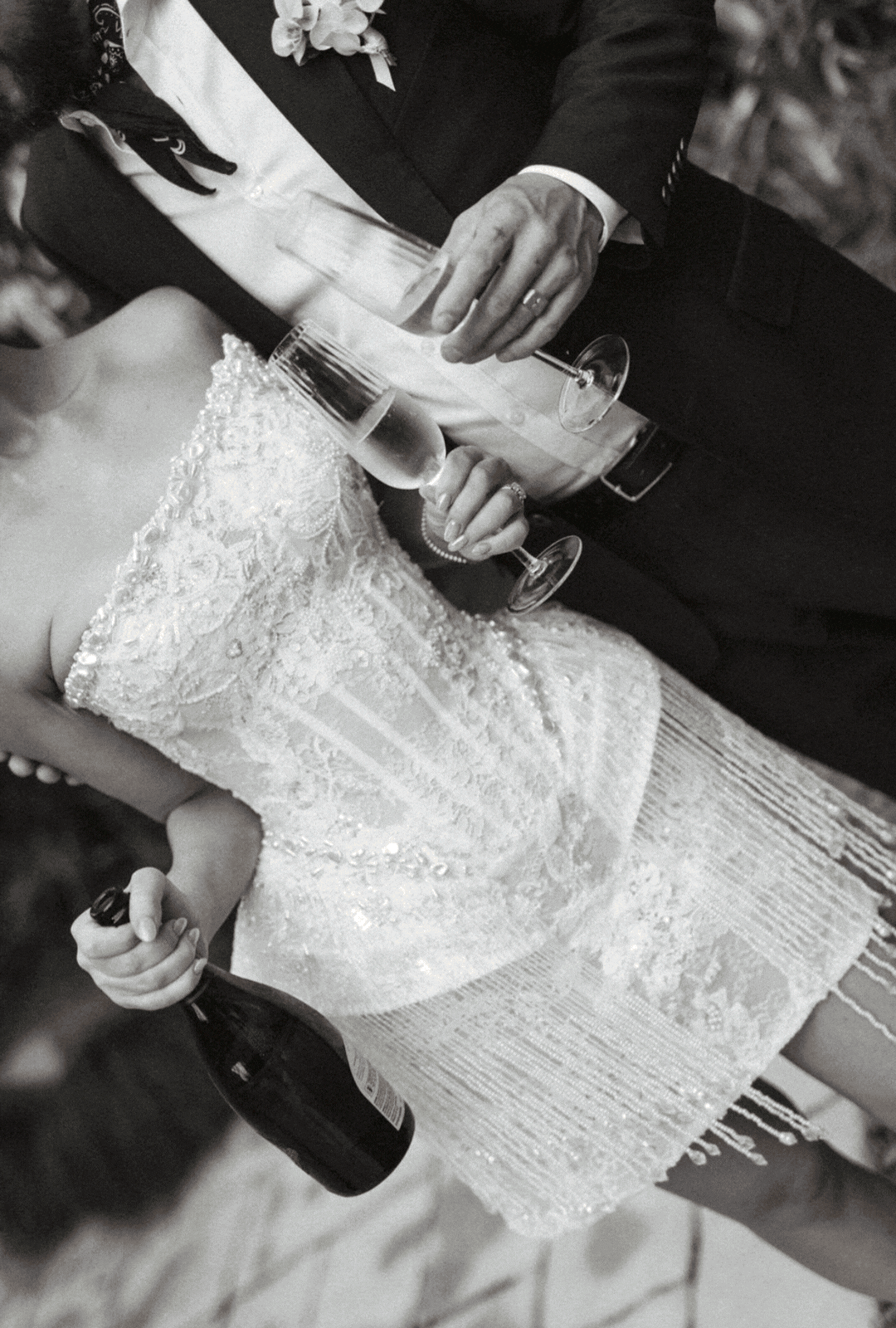 A bride and groom at a wedding celebration, holding glasses of champagne, with the bride in a detailed lace wedding dress and the groom in a dark suit. The groom is also holding a bottle of beer.