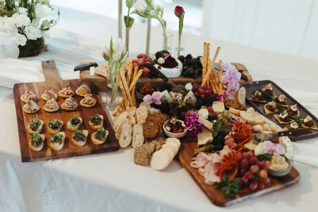A cheese and charcuterie board with various cheeses, sliced meats, crackers, bread, grapes, berries, and edible flowers, arranged on wooden serving boards with small bowls of accompaniments.