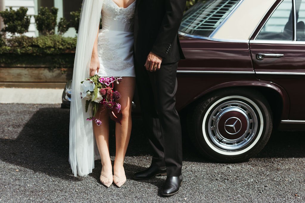 A bride and groom standing next to a vintage maroon Mercedes-Benz car, with the bride holding a bouquet of flowers.