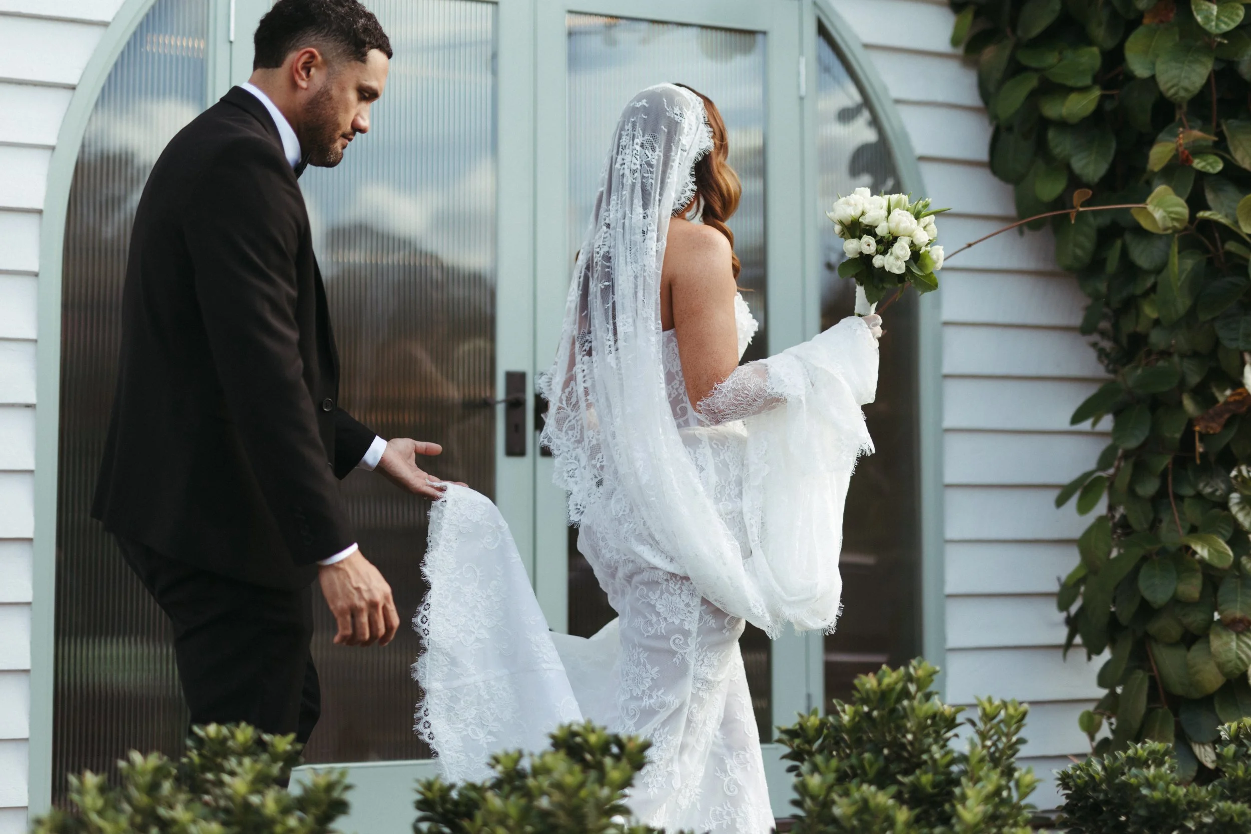 A couple dressed in wedding attire walking outside, with the bride in a white lace wedding gown and veil holding a bouquet of white flowers, and the groom in a black suit. The scene features a white house with green foliage.