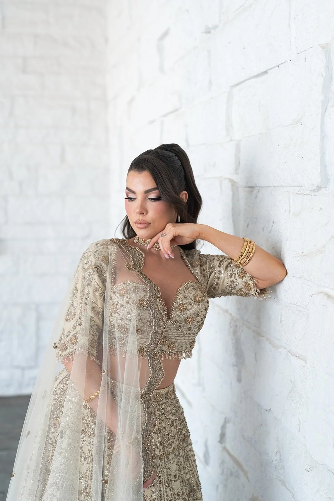 A woman dressed in a white wedding gown, with dark brown hair styled in loose waves and makeup, looking at the camera while holding a sheer veil.
