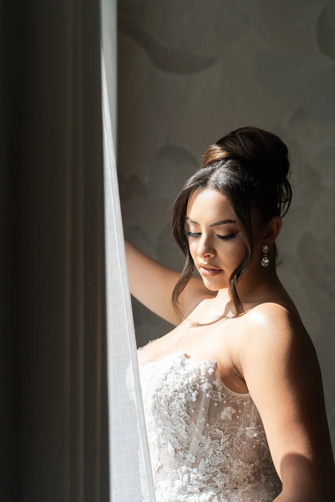 A woman in a white lace wedding dress with long sheer sleeves, standing in front of a blue patterned wall. She has long dark hair styled in loose waves and is wearing a bridal veil.