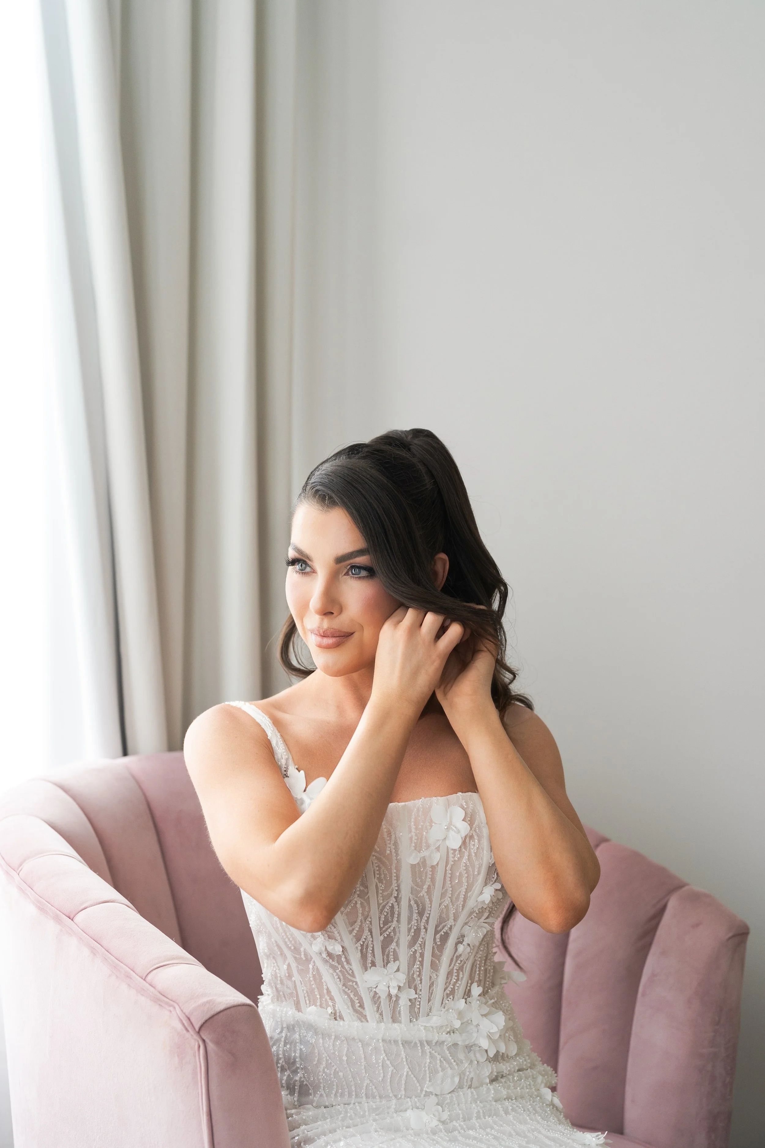 A woman in a wedding dress with lace detailing, wearing large earrings, holding a white veil close to her face, with dark hair styled in loose waves, posing against a plain white background.