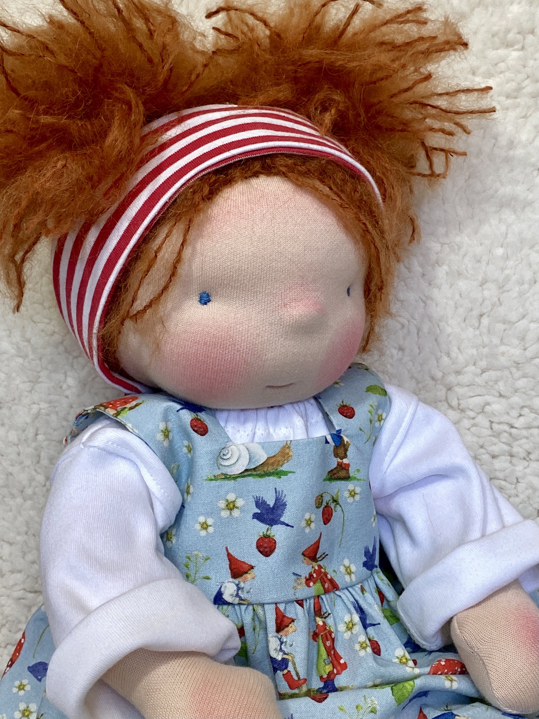Waldorf doll with red hair and blue eyes wearing a striped headband and light blue overalls.