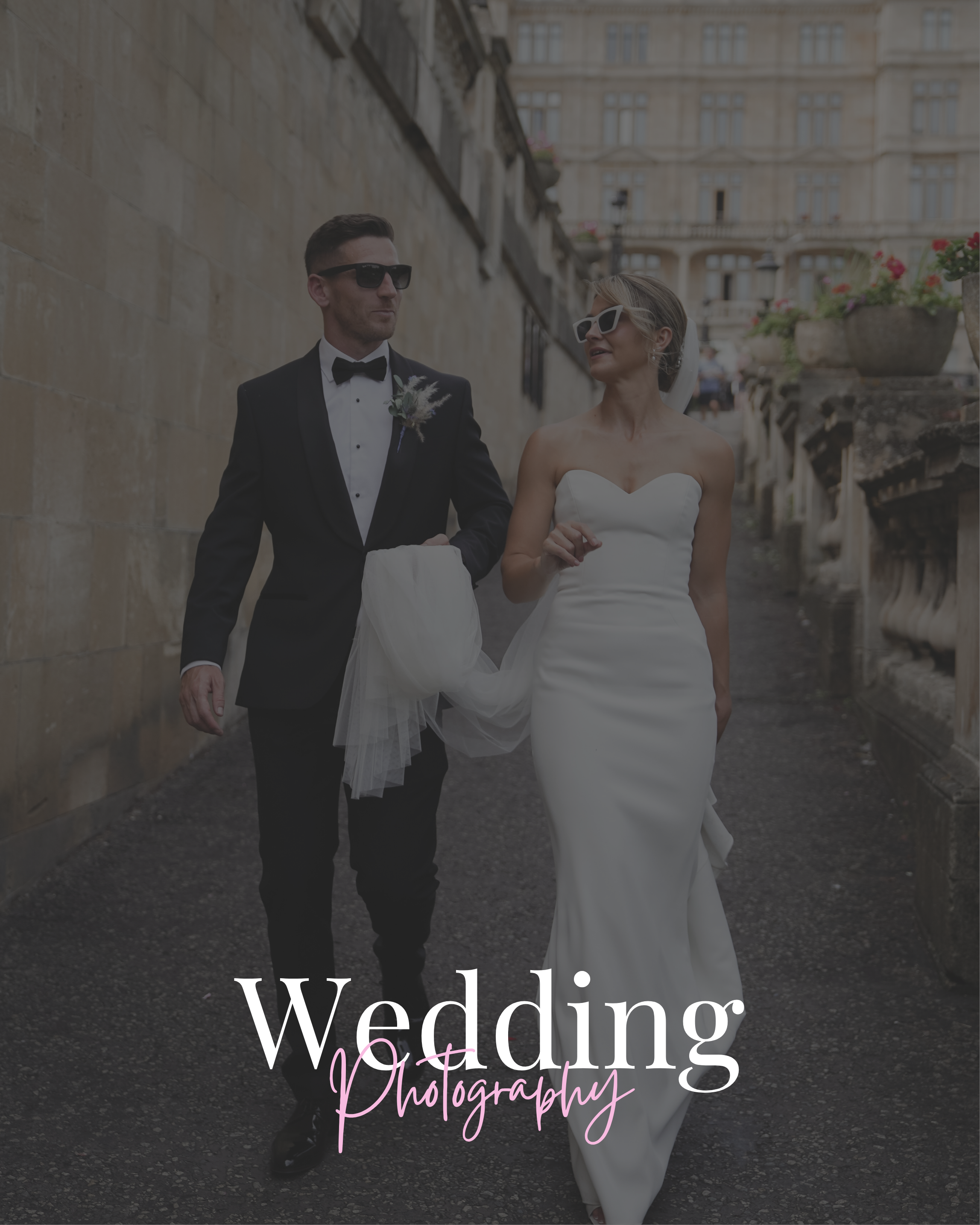 A bride and groom walking on a city street. The groom is wearing a black tuxedo with a bow tie and sunglasses, holding the bride's veil. The bride is wearing a strapless white wedding gown and white sunglasses.
