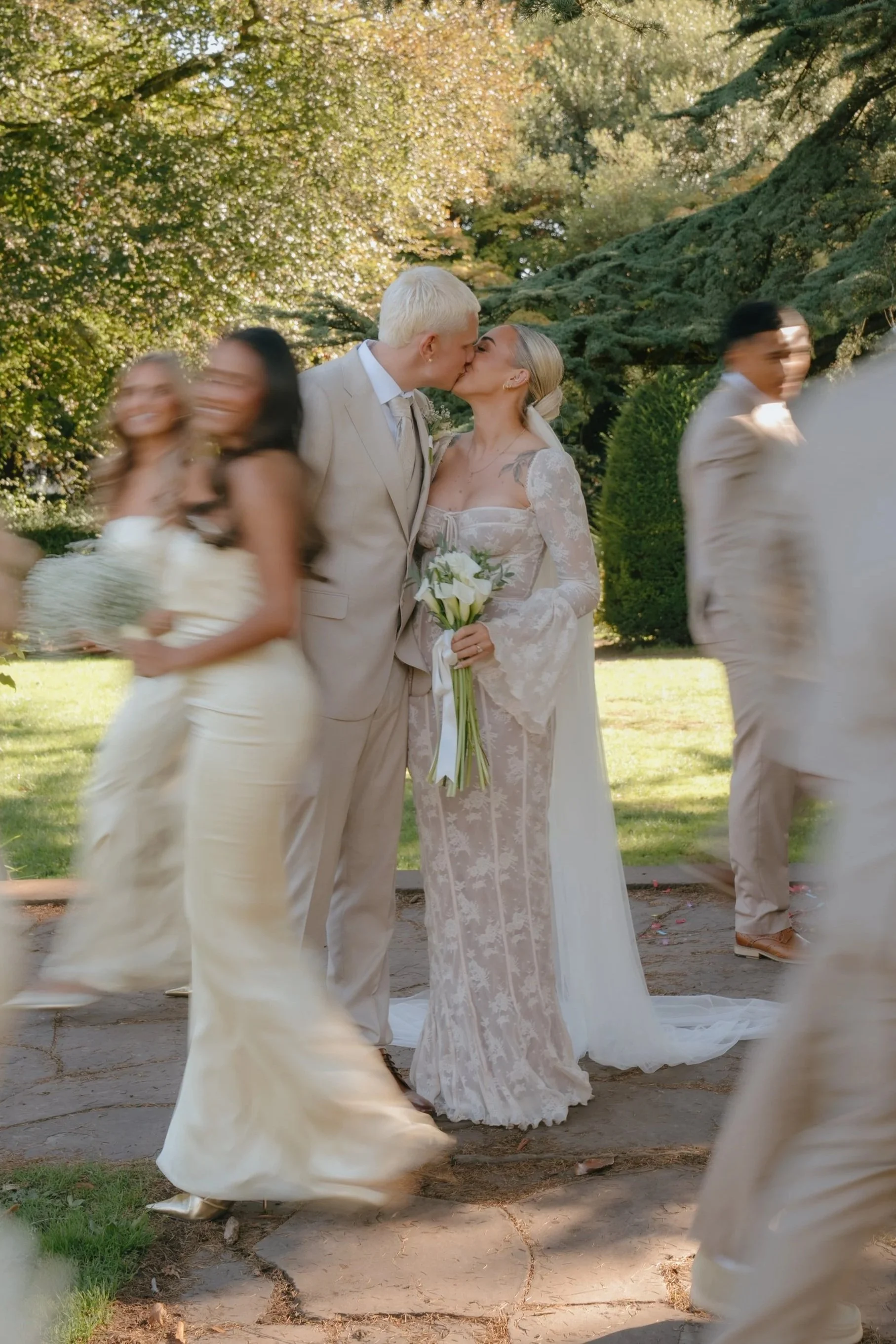 A bride and groom kiss outdoors during their wedding ceremony, surrounded by blurred wedding guests in beige attire and green trees in the background.