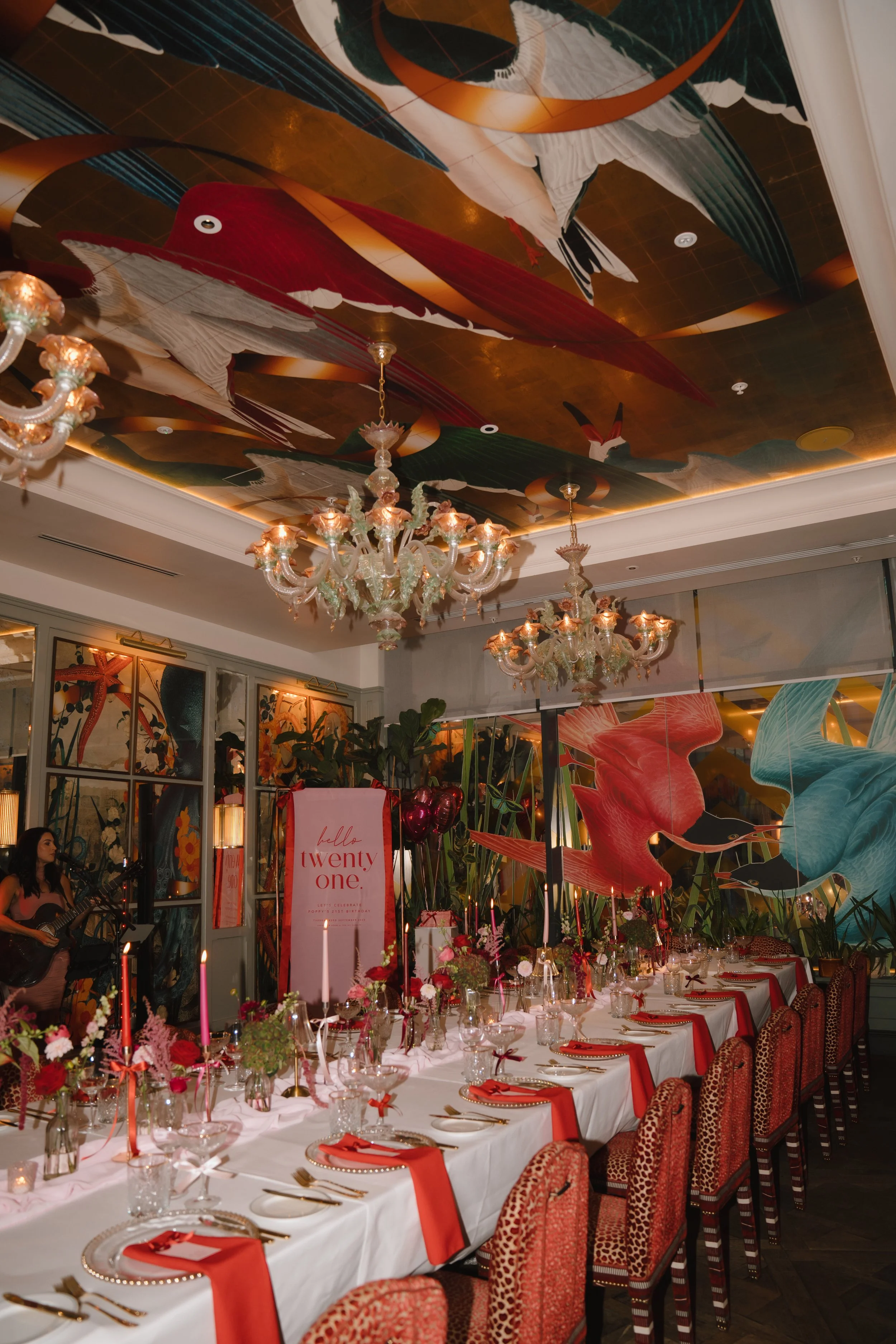 A decorated dining room set for a celebration, with a long table covered with white tablecloths, red napkins, and floral centerpieces. Elegant chandeliers hang from a ceiling adorned with large, colorful bird murals, and there are tall plants and bal
