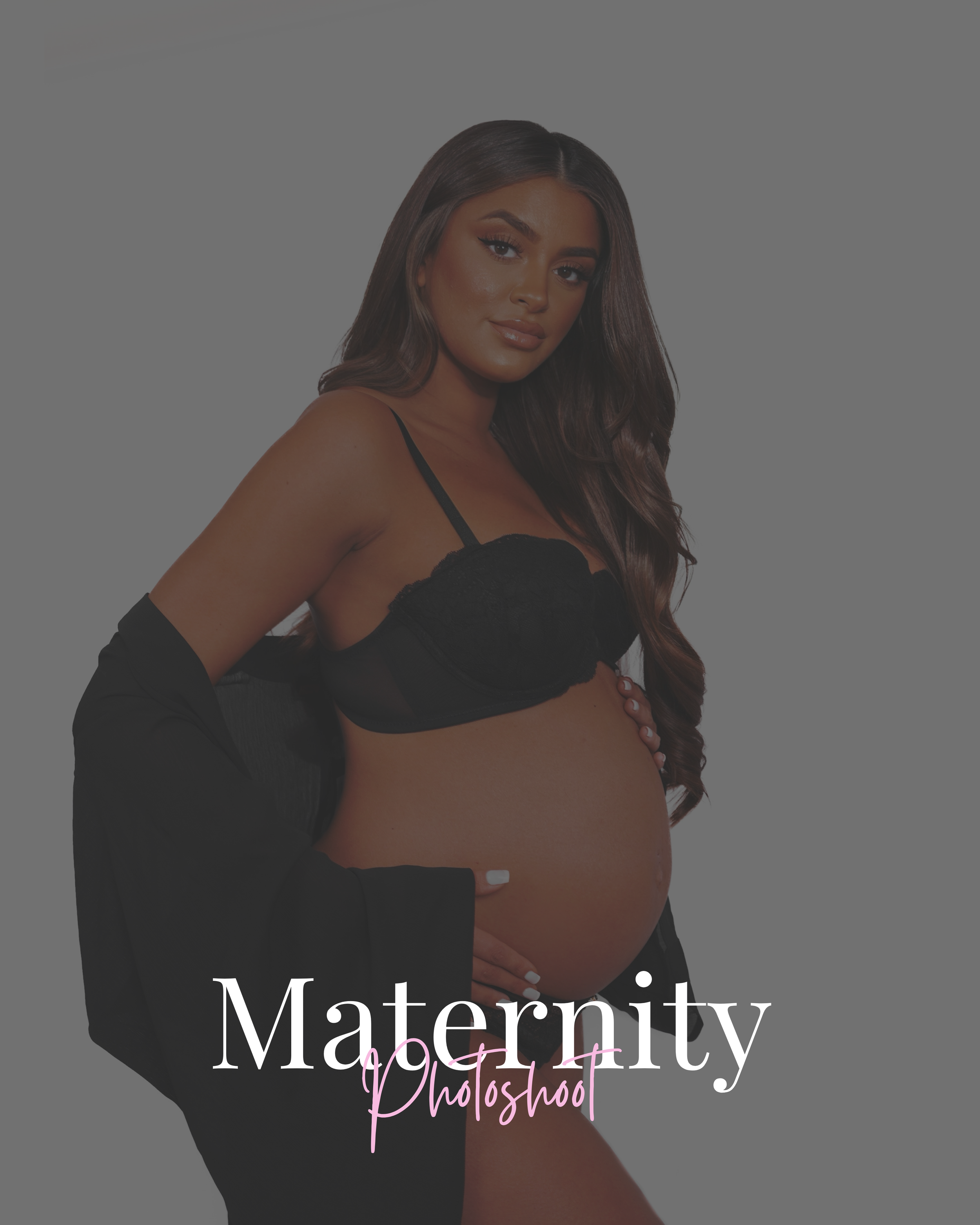 A pregnant woman with long wavy hair, standing against a gray background, wearing black lingerie and a black jacket, with her hand on her belly, looking at the camera.
