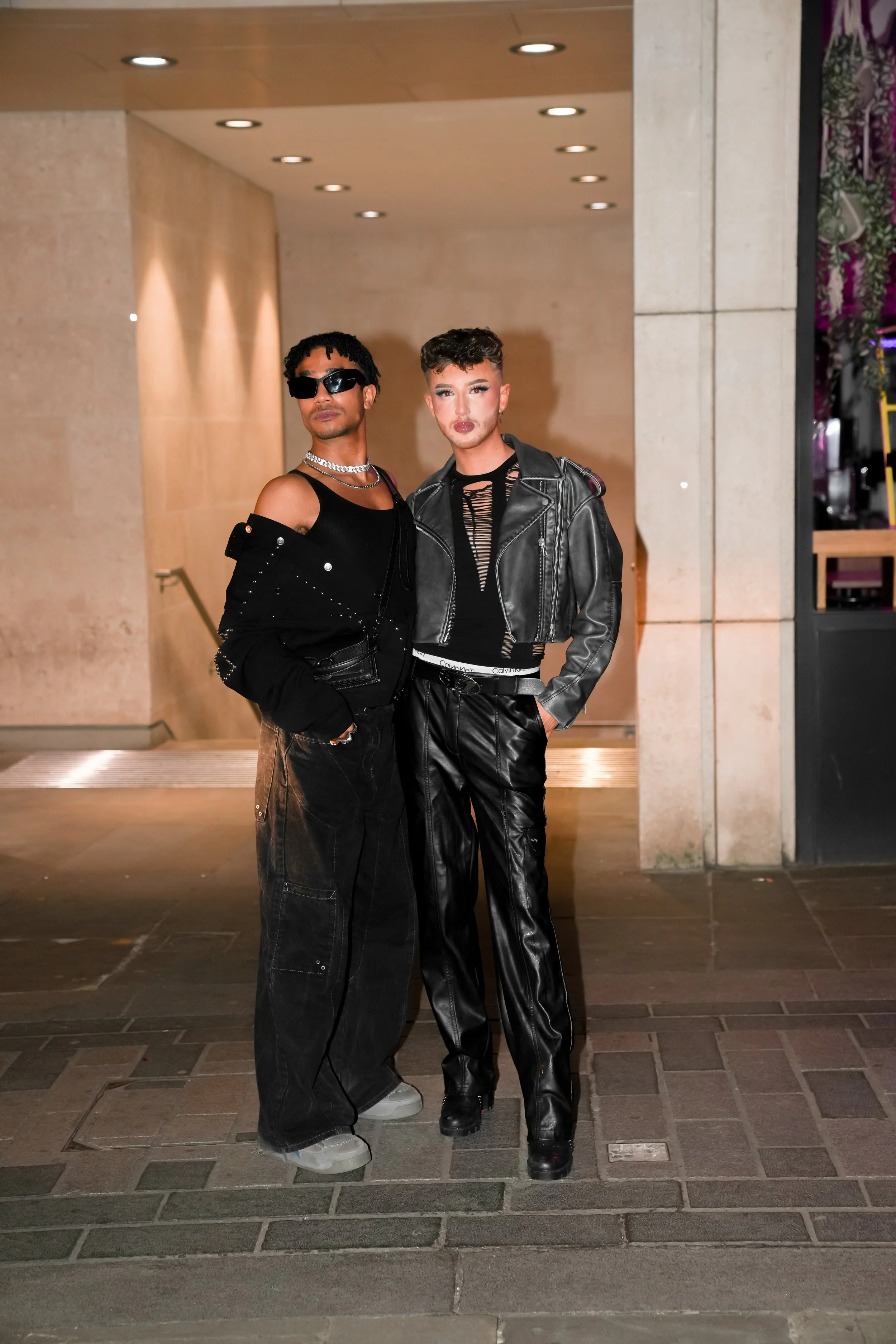 Two individuals standing together in an urban setting at night, dressed in fashionable, edgy outfits with leather and black styles.