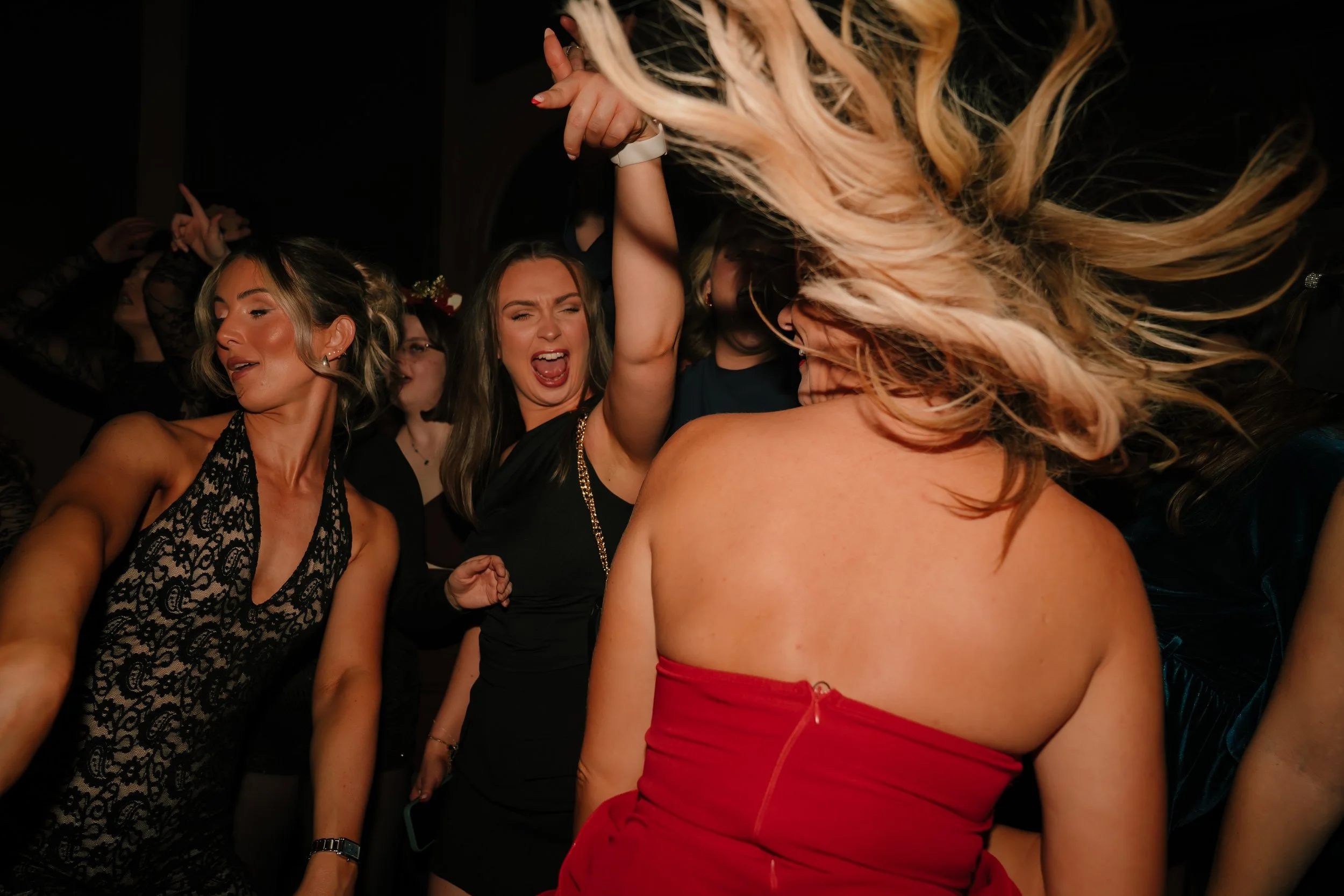Group of women dancing and enjoying themselves at a party or celebration, with one woman in a red strapless dress in the foreground, and others wearing black dresses.