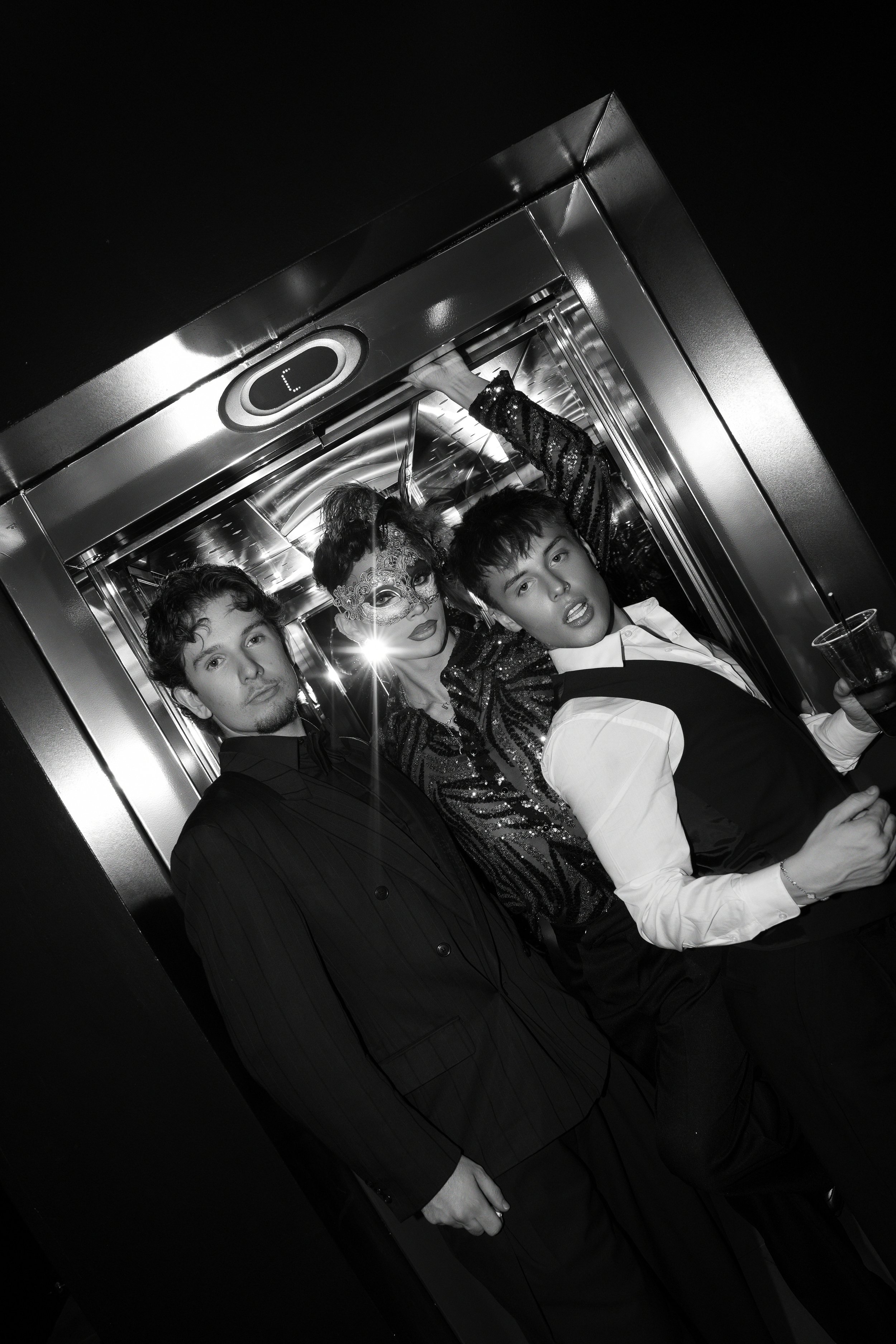 Three people, two men and one woman, in formal attire inside an elevator, with the woman wearing a masquerade mask and sequined jacket, one man holding a drink, all posing for a photo.