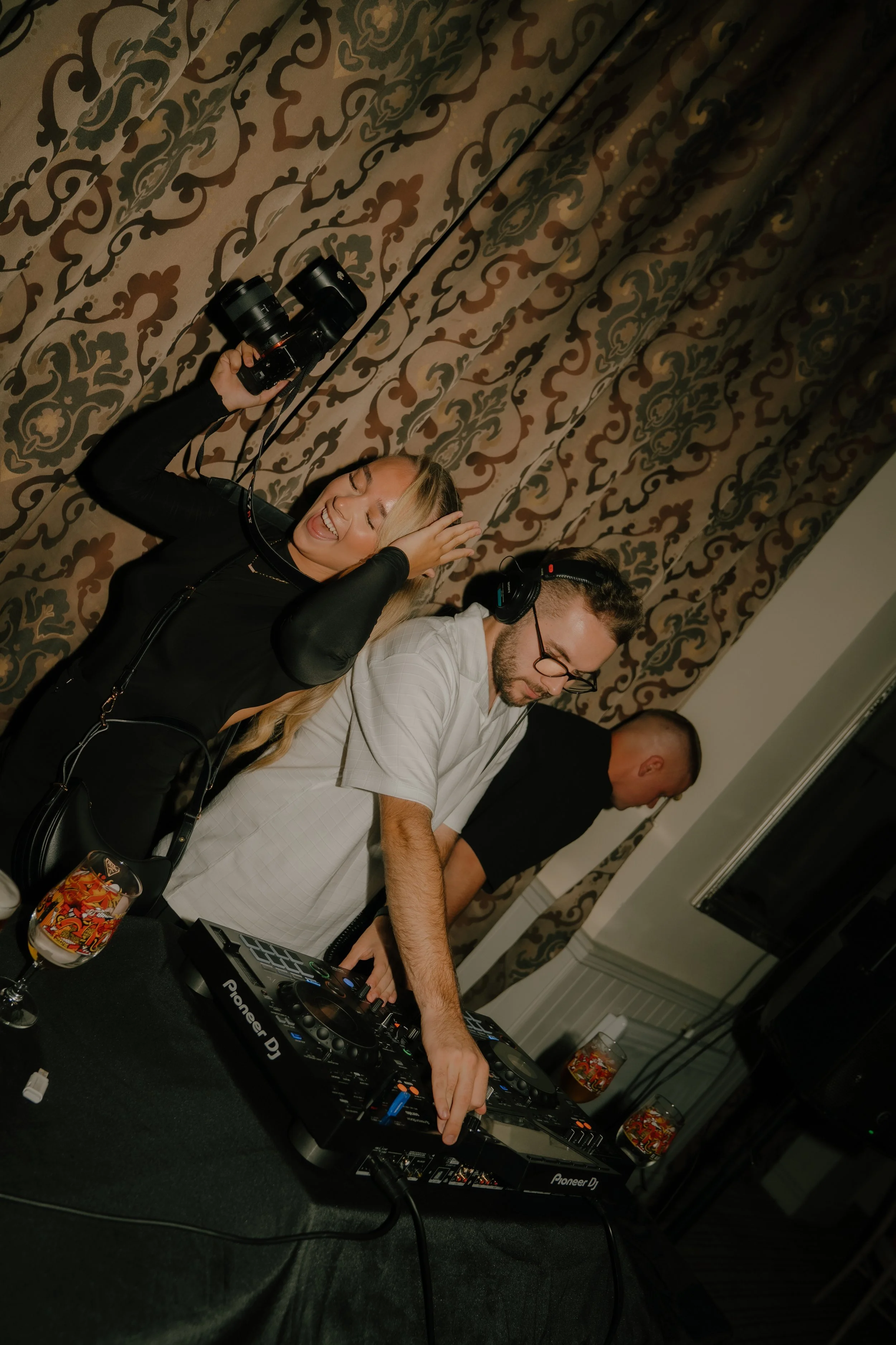 Two DJs, a woman and a man, are playing music at an event. The woman is holding a camera and smiling, while the man is focusing on the DJ equipment. A third person is visible in the background.