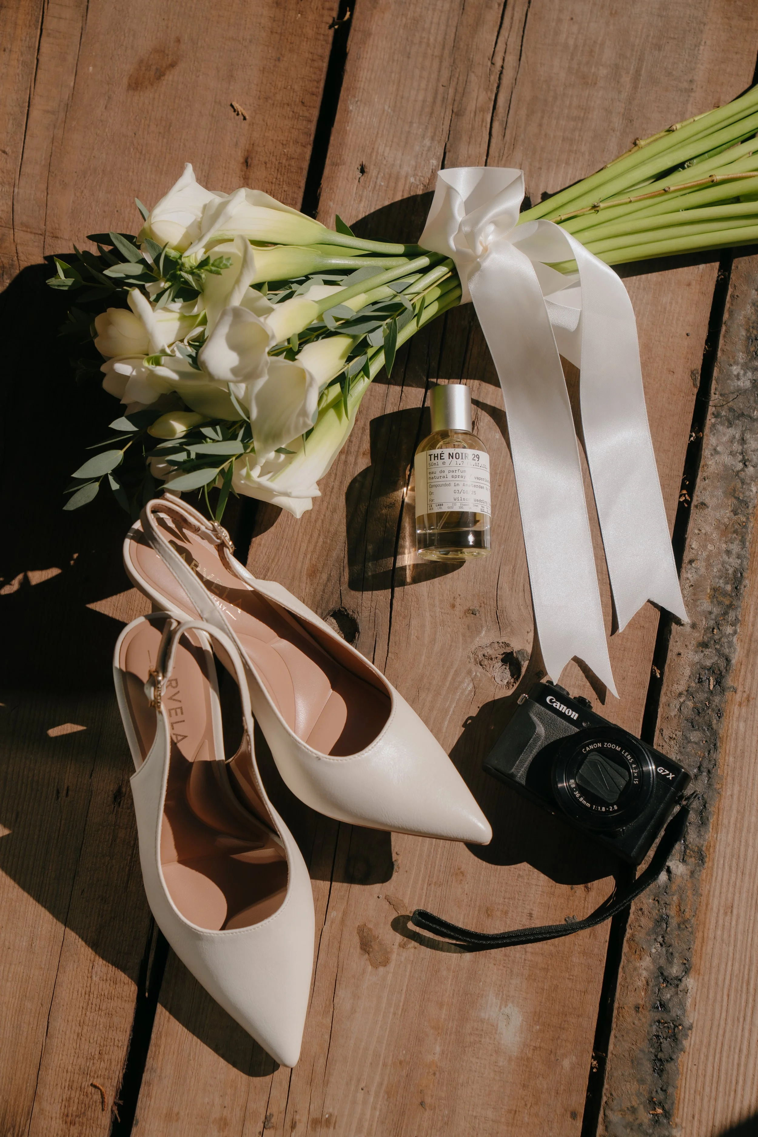 A bridal bouquet of white calla lilies with green leaves, a pair of white high-heeled shoes, a small bottle of perfume, a vintage camera, all on a wooden surface.