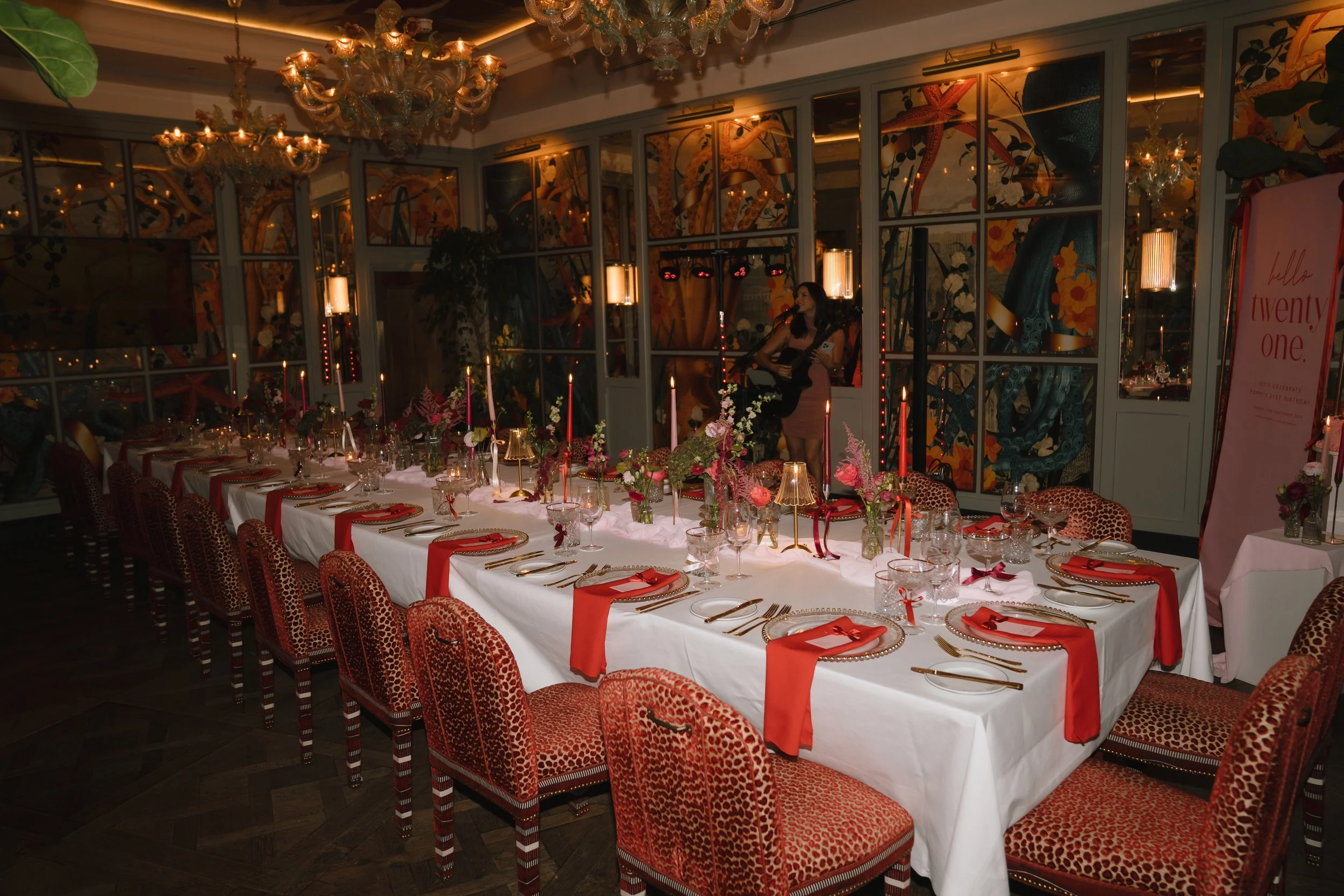 A long dining table set for a celebration with red and white accents, surrounded by leopard print chairs, lit candles, floral centerpieces, and ornate chandeliers in a decorated indoor space.