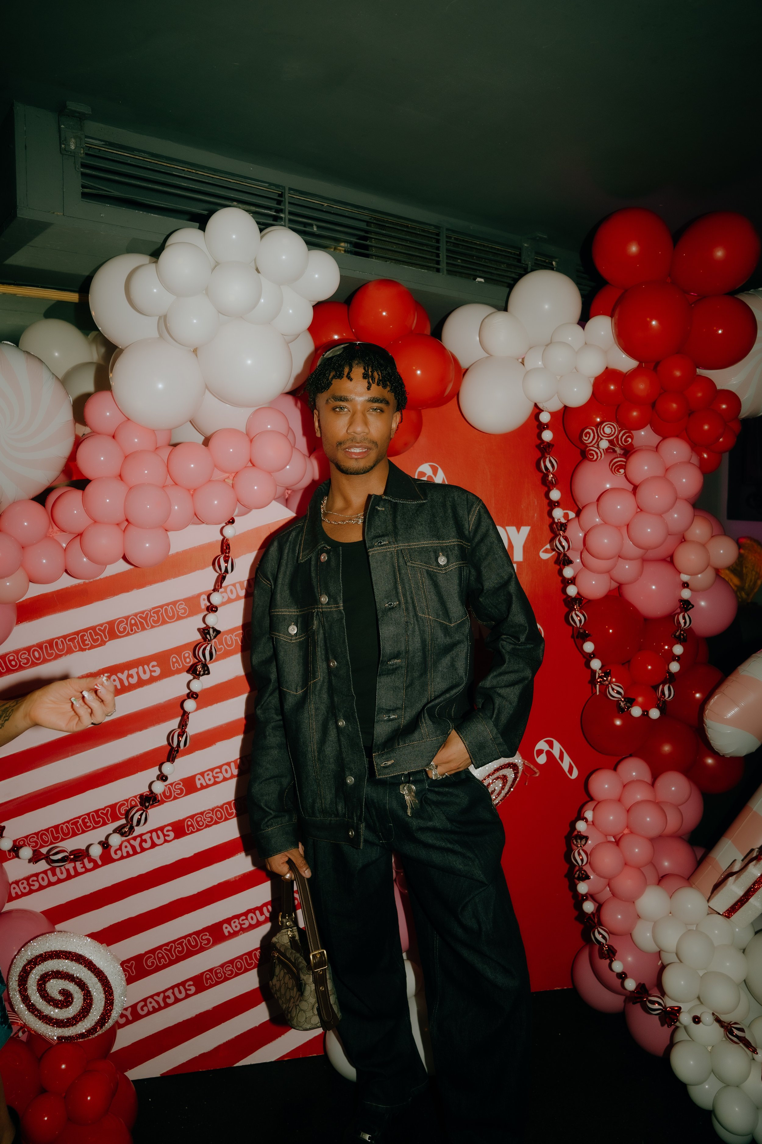 A young man with short curly hair, wearing a black shirt and a dark denim jacket, standing in front of a festive backdrop with balloons and a red and white theme.