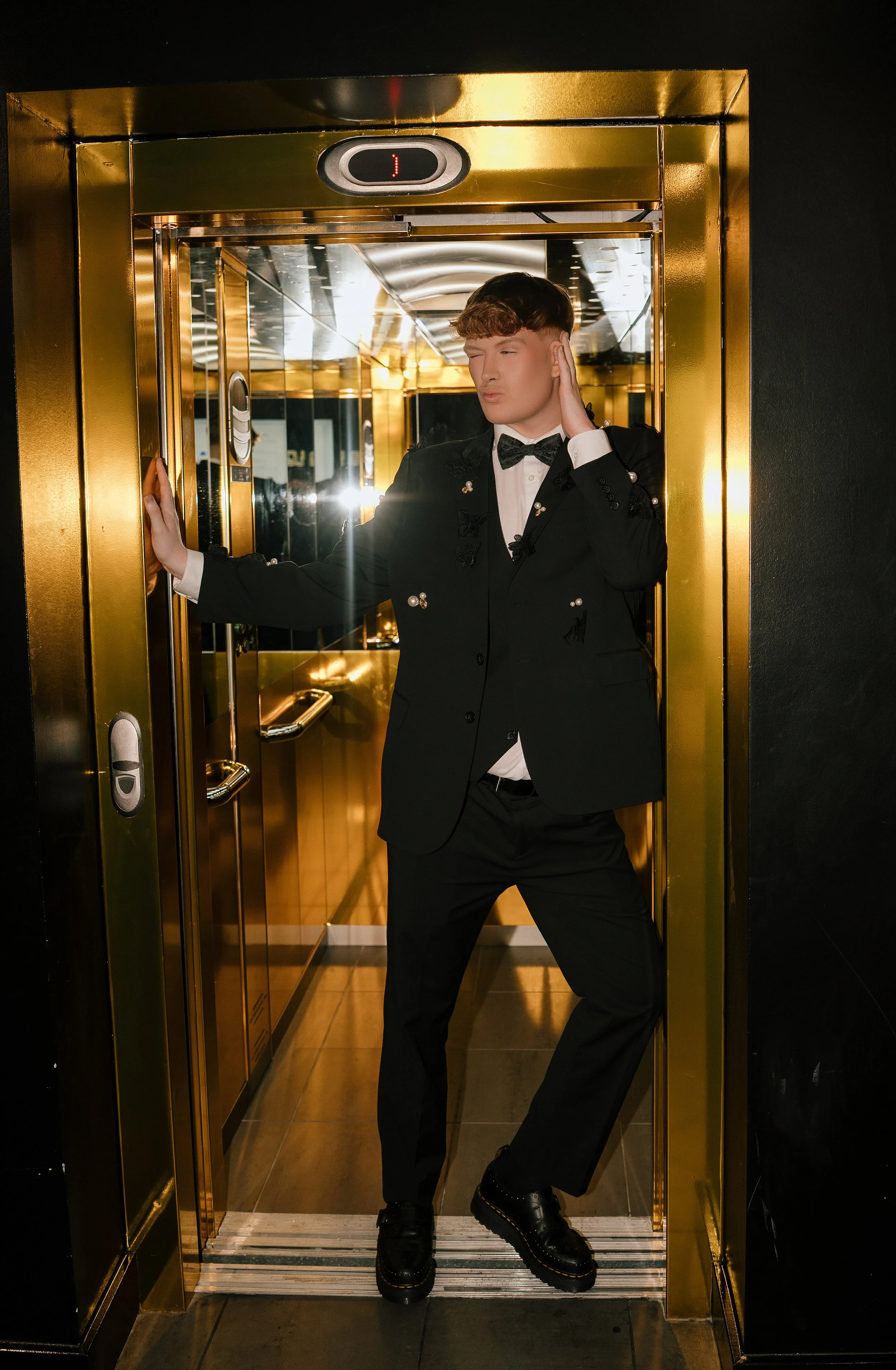 A young man in a tuxedo posing inside a gold-colored elevator with mirrored walls.